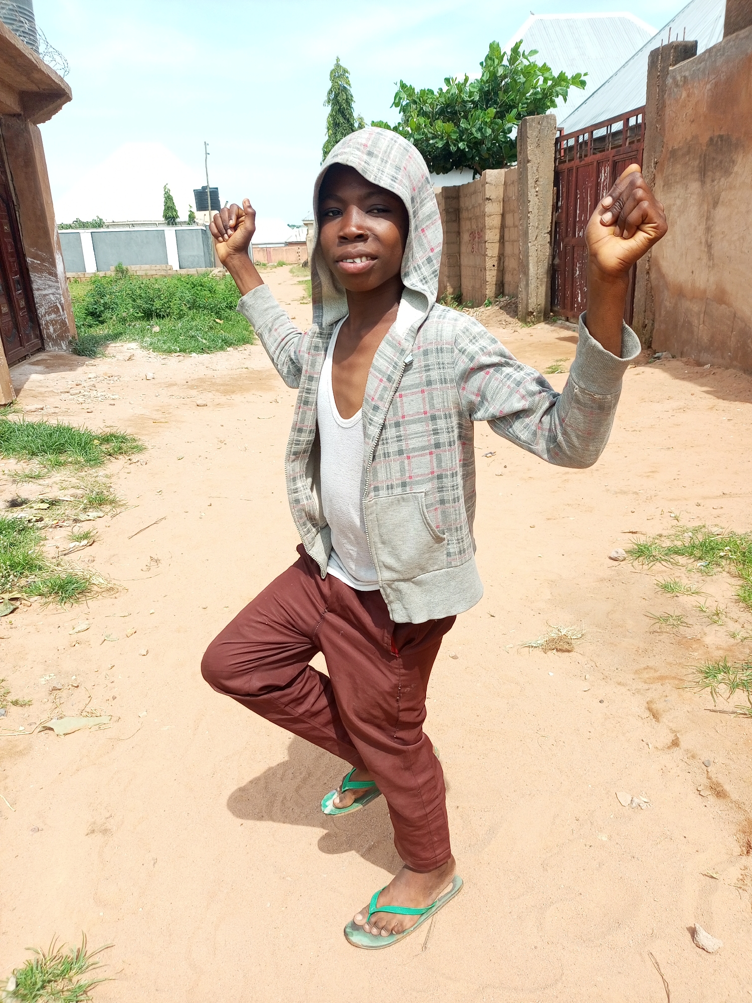 A young boy is standing in a dusty, unpaved area, likely a street or yard, in Tempure, Nigeria. He is wearing a grey hooded jacket with a checkered pattern over a white tank top and maroon pants. His left leg is bent, and he is balancing on his right foot. Both of his hands are raised, with his fists clenched. The background consists of rustic buildings, a metal gate, and some vegetation under a clear sky. The ground is sandy and uneven, with scattered debris.