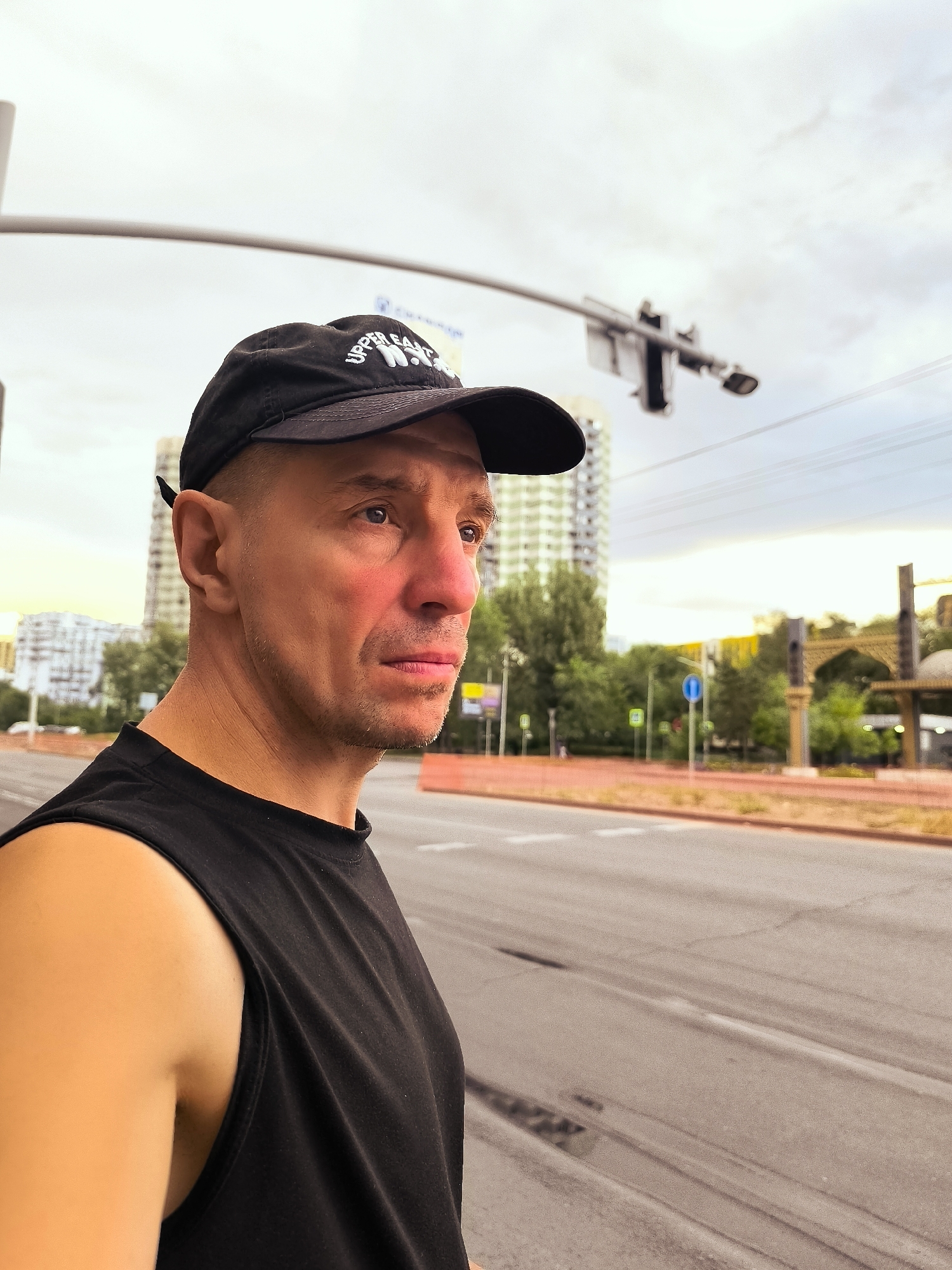 A man wearing a black baseball cap and a black sleeveless t-shirt is standing on a city street in Almaty, Kazakhstan. He is looking to his right, with buildings, trees, and a traffic light visible in the background. The sky is overcast. The man's facial features are distinct: he has a fair complexion, a short haircut, and appears to be in his 40s or 50s. The background shows a typical urban landscape with a multi-lane road. The man is positioned in the foreground, with the city scene serving as a backdrop, suggesting he might be a pedestrian waiting to cross the street or simply observing his surroundings.
