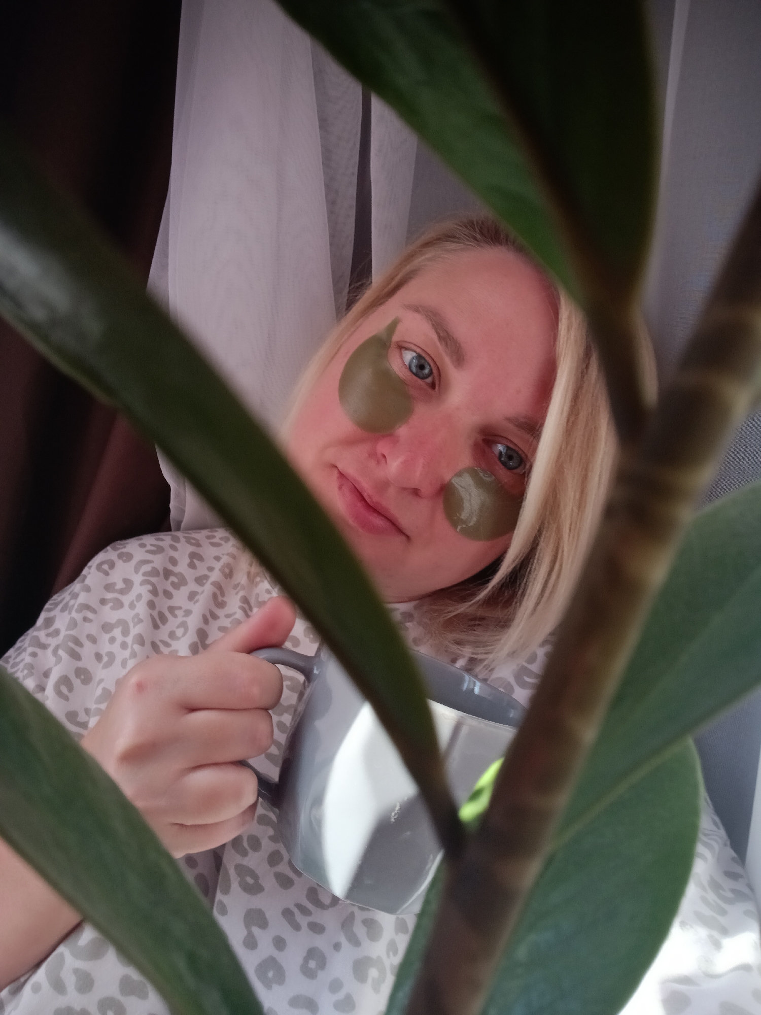A light-skinned woman with blonde hair is holding a grey mug while wearing green under-eye patches. She is positioned behind a potted plant with green leaves. The background includes a window with sheer white curtains and a dark brown curtain. The location is Chaikovsky, Russia.