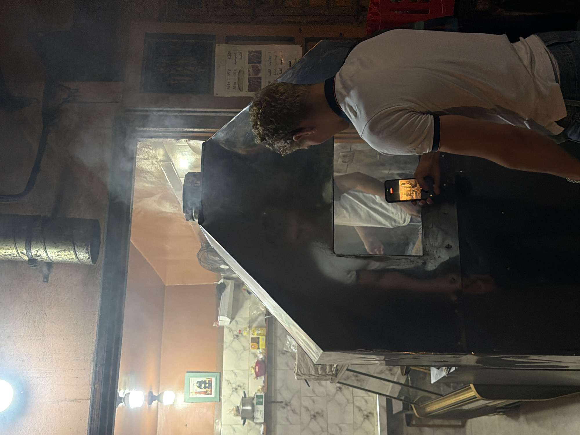 In Marrakesh, Morocco, a young man with curly blond hair is standing in front of a large, black, angular structure that appears to be a food stall or oven, emitting steam. He is holding a smartphone in his right hand, angled to capture a video of himself and the structure. He is wearing a white polo shirt with a dark collar and blue trim on the sleeves, and blue jeans. The background shows a kitchen-like environment with tiled walls, shelves holding food items and utensils, and a framed picture on the wall. The lighting is dim, with a warm glow emanating from overhead lamps. The overall scene suggests a casual, possibly touristy, experience in a food establishment.