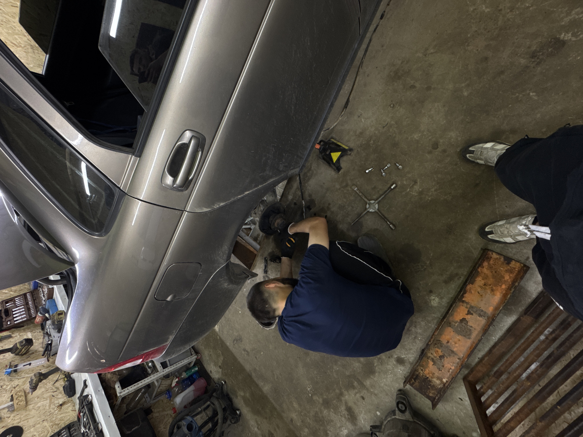 The image depicts an indoor car repair workshop located in Perm, Russia. A single individual is observed from a high angle, crouched on a stained concrete floor, actively performing maintenance on the rear left side of a grey sedan. The person wears a dark blue short-sleeved t-shirt, dark pants with a white stripe, grey athletic shoes, and dark work gloves, with dark hair tied back. The car's trunk is open, and its rear left wheel has been removed, exposing the wheel hub and brake assembly. A yellow and black jack stand is positioned beneath the vehicle near the rear passenger door for support. On the concrete floor directly in front of the worker are a four-way lug wrench and several loose lug nuts. The car's exterior panels are dusty, and a minor blemish or dent is visible above the driver's side rear taillight. A man's face, holding a smartphone, is clearly reflected in the car's rear passenger window, indicating the photographer's presence. In the immediate foreground, the feet of the photographer, clad in dark pants and white and grey athletic shoes, are visible alongside two rusted metal objects: a flat, orange-brown beam and a parallel-barred grate. The background displays a workshop environment with wood-paneled walls, on which various tools such as power drills, wrenches, and toolboxes are neatly hung or stored.