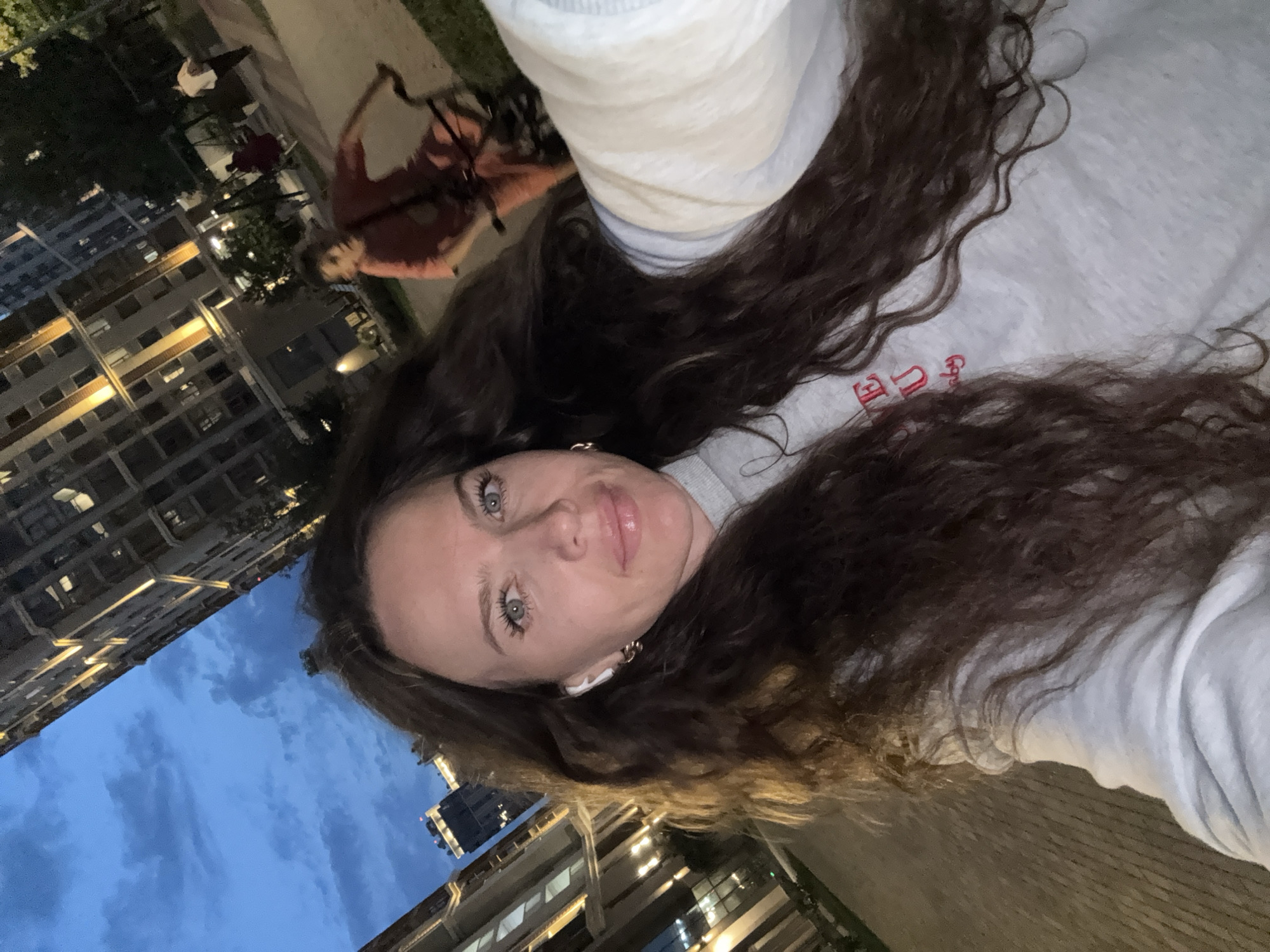 The image shows a young woman taking a selfie outdoors in the evening. She has long, dark, wavy hair and is wearing a grey hoodie with red lettering. She has earbuds in and is wearing hoop earrings. In the background, there are modern, tall buildings with illuminated windows, typical of a city environment. To the right, a person is riding a bicycle. The sky is a deep blue, indicating dusk or early night. The ground appears to be paved. The overall impression is that of a person enjoying a leisurely evening in a city setting, possibly Moscow, Russia, given the architecture and the general urban atmosphere.