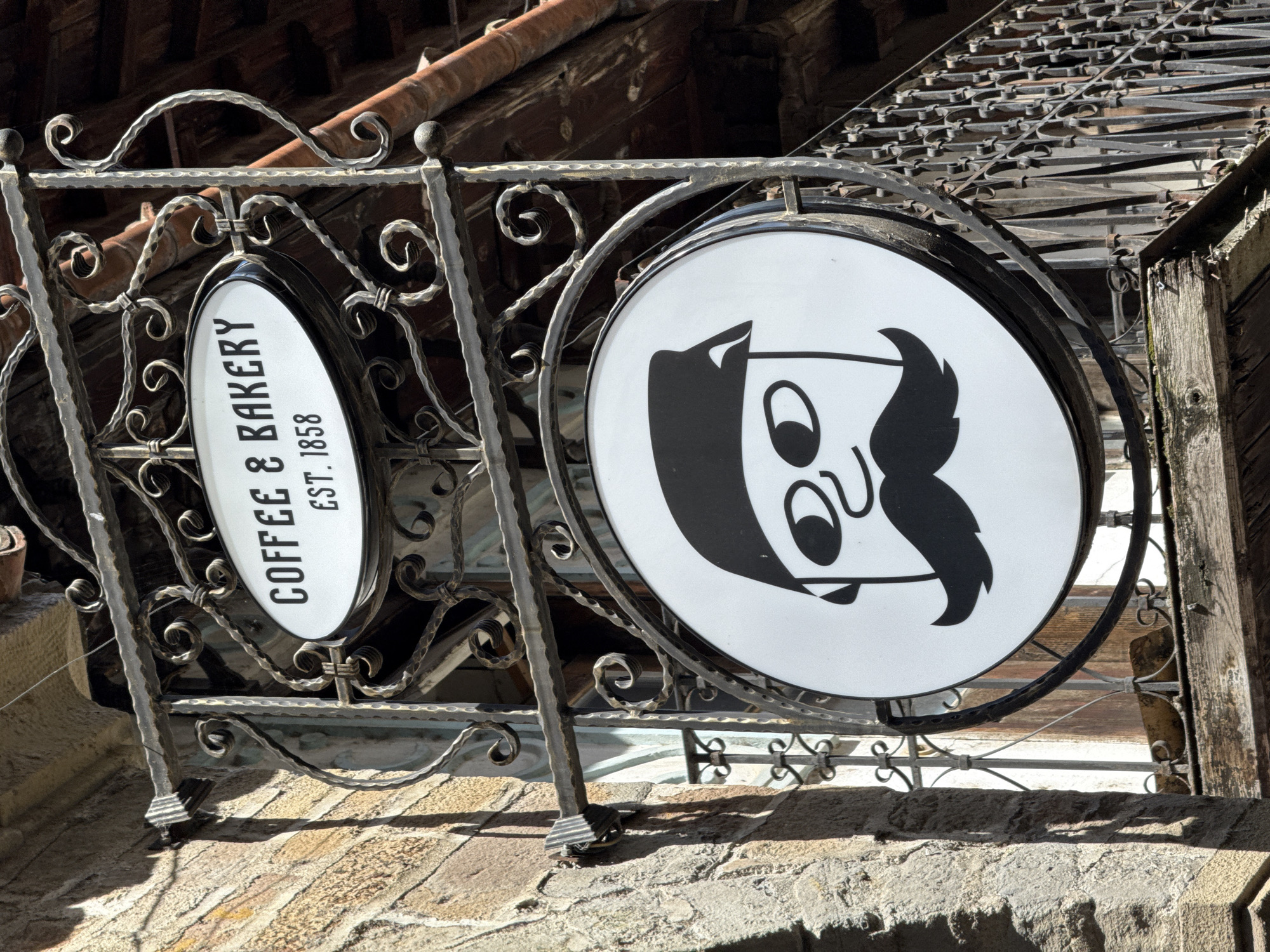 The image captures two signs for a coffee and bakery establishment, likely located in Veliko Tarnovo, Bulgaria. The signs are mounted on an ornate, wrought-iron railing. The sign on the left is oval-shaped and reads "COFFEE & BAKERY EST. 1858". The sign on the right is circular and features a stylized, cartoon-like drawing of a man's face with a mustache. The wrought-iron railing is intricately designed with curls and decorative elements. The signs appear to be attached to a building with a brick or stone facade. In the background, there is a wooden awning or structure with iron grates, suggesting an older building. The scene is bathed in sunlight, casting shadows on the wall and signs.