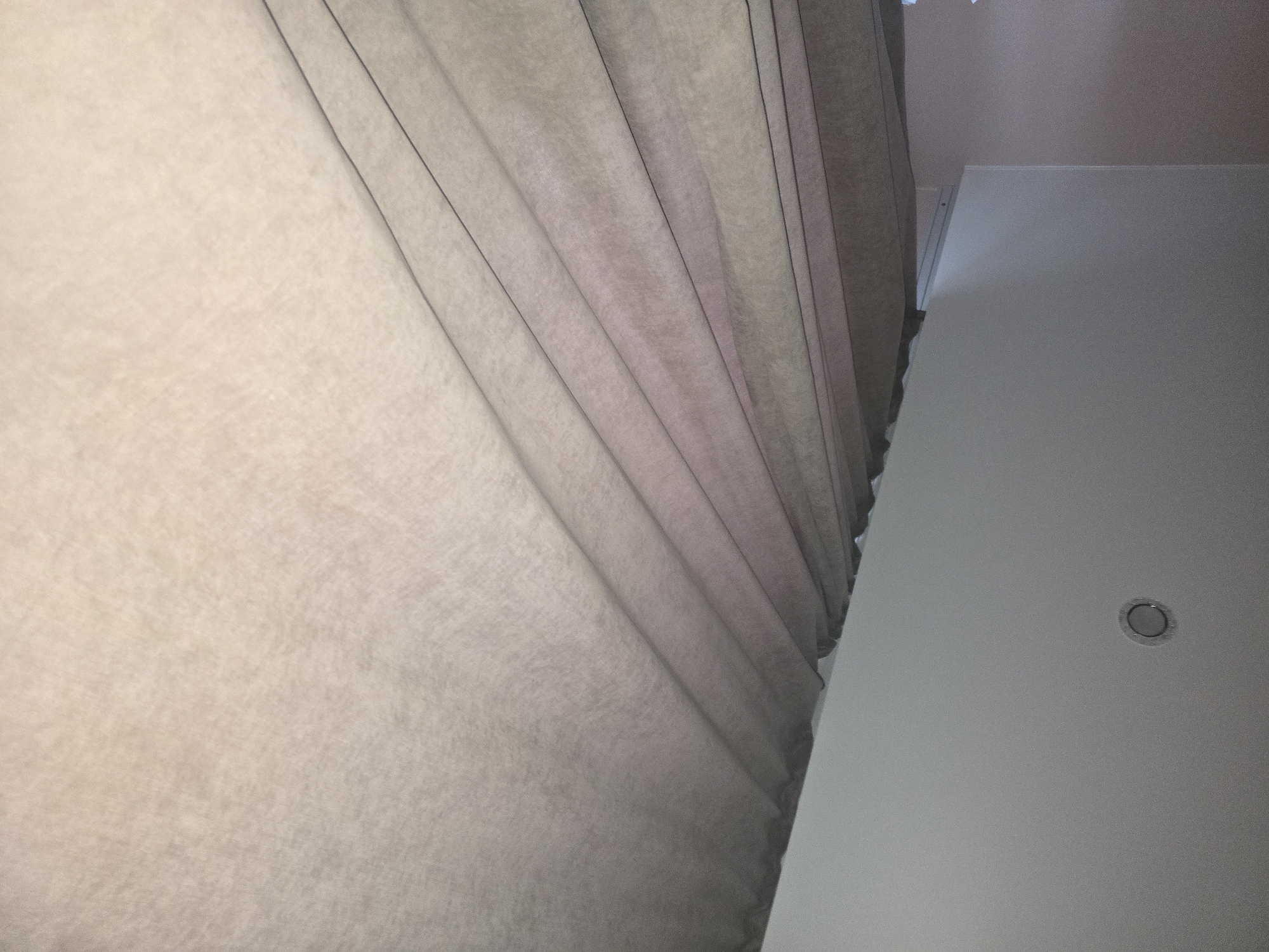 The image shows a ceiling with a recessed light fixture, partially obscured by draped sheer curtains. The curtains are light brown and hang in vertical folds, allowing some light to filter through. The ceiling is white and appears to be a standard residential or office space. The photograph is taken from a low angle, looking upwards. There are no people or animals visible in the image, nor are there any specific landmarks that would indicate a location in Ulan-Ude, Russia. The scene is static and lacks any discernible action or interaction. The overall environment is neutral and domestic.