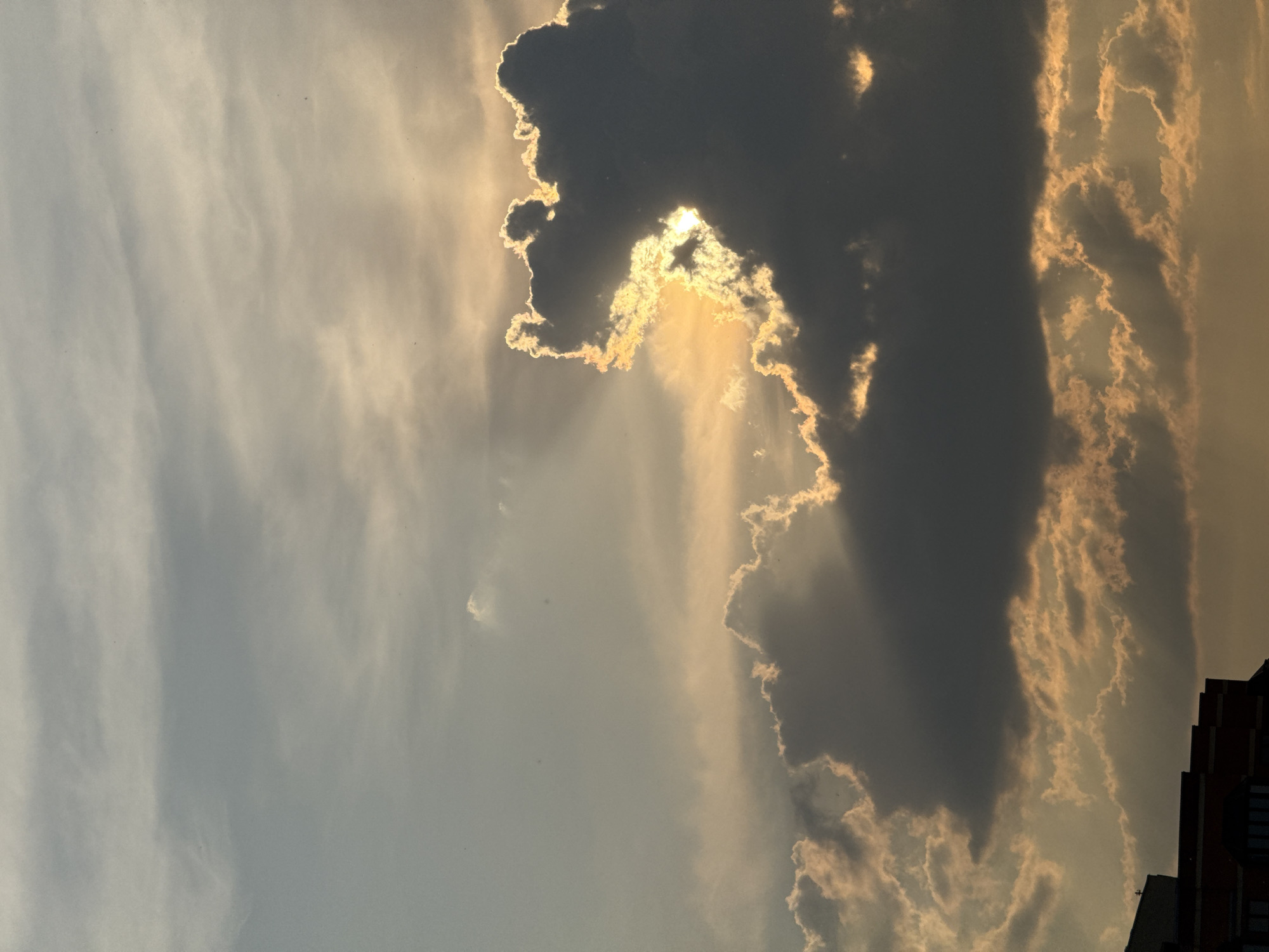 The image shows a dramatic sky with clouds and sunlight. The sun is partially obscured by a large, dark cloud in the center right of the frame, with rays of light peeking through its edges, illuminating thinner, wispy clouds with a golden hue. The sky transitions from a lighter, grayish-blue at the top to warmer, orange and yellow tones closer to the horizon. In the bottom left corner, the silhouette of a building with red brick and dark windows is visible, suggesting an urban setting. The overall impression is of a sunset or sunrise, with the interplay of light and shadow creating a powerful visual effect. No people or specific actions are depicted. The image does not contain any identifiable landmarks or features that would definitively place it in Moscow, Russia.