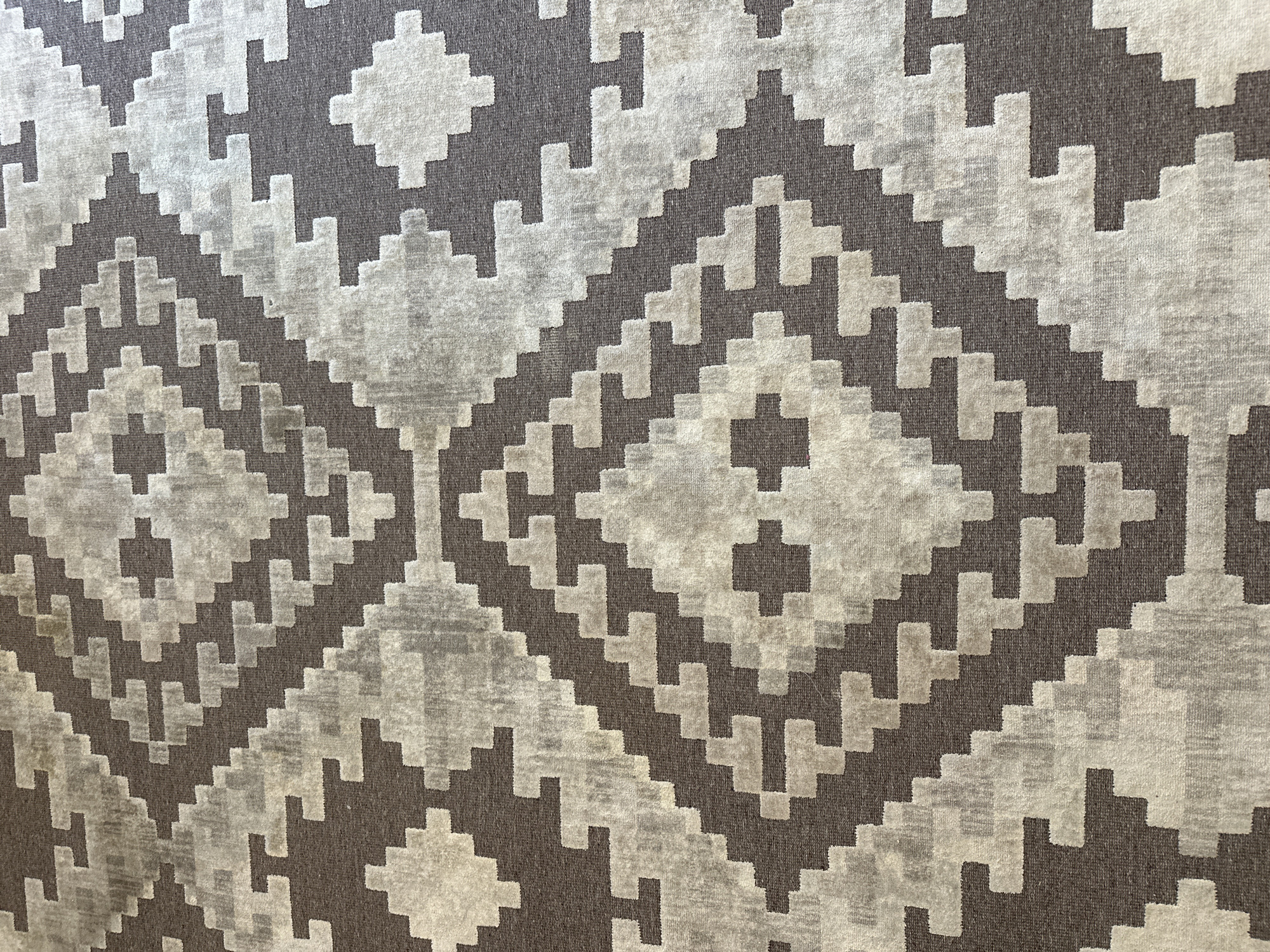 The image is a close-up, overhead view of a rug with a geometric diamond pattern in shades of brown and grey. The pattern consists of repeated angular shapes creating a tessellating effect across the surface of the rug. The rug appears to be indoors, likely on a floor. There is no discernible information in the image that would suggest a location in Minsk, Belarus.