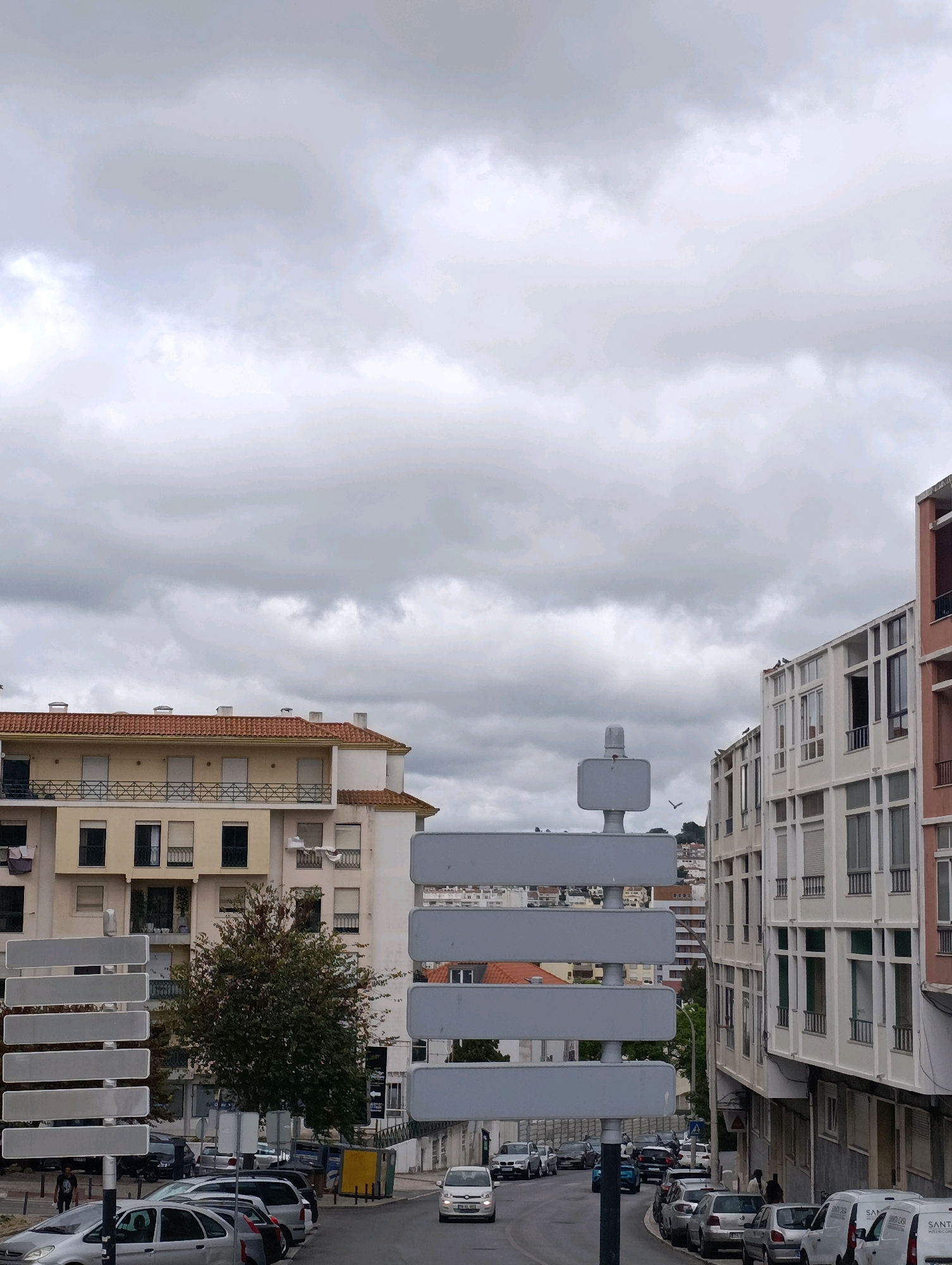 This is a photograph taken from a street level perspective, looking upwards towards a cloudy sky and several buildings. In the foreground, a grey traffic signpost with multiple rectangular signs is visible on the left. The signs are blank. Cars are parked along the street on the left and right. A person is walking on the sidewalk on the left side of the street. A white car is driving down the street in the middle of the image. On the left side of the image, there is a building with a red-tiled roof and cream-colored walls, with balconies. On the right side, there are modern apartment buildings with many windows and balconies. In the background, more buildings are visible on a hill, and trees are interspersed among them. The overall impression is an urban street scene under an overcast sky. The signs and buildings suggest this is a public area within a city. The presence of parked and moving vehicles, pedestrian activity, and residential and commercial buildings indicates an urban environment. The architectural style of the buildings, particularly the modern apartment blocks with their repetitive window patterns, is characteristic of many European cities. The specific reference to Setúbal, Portugal, suggests this scene is located within that city.