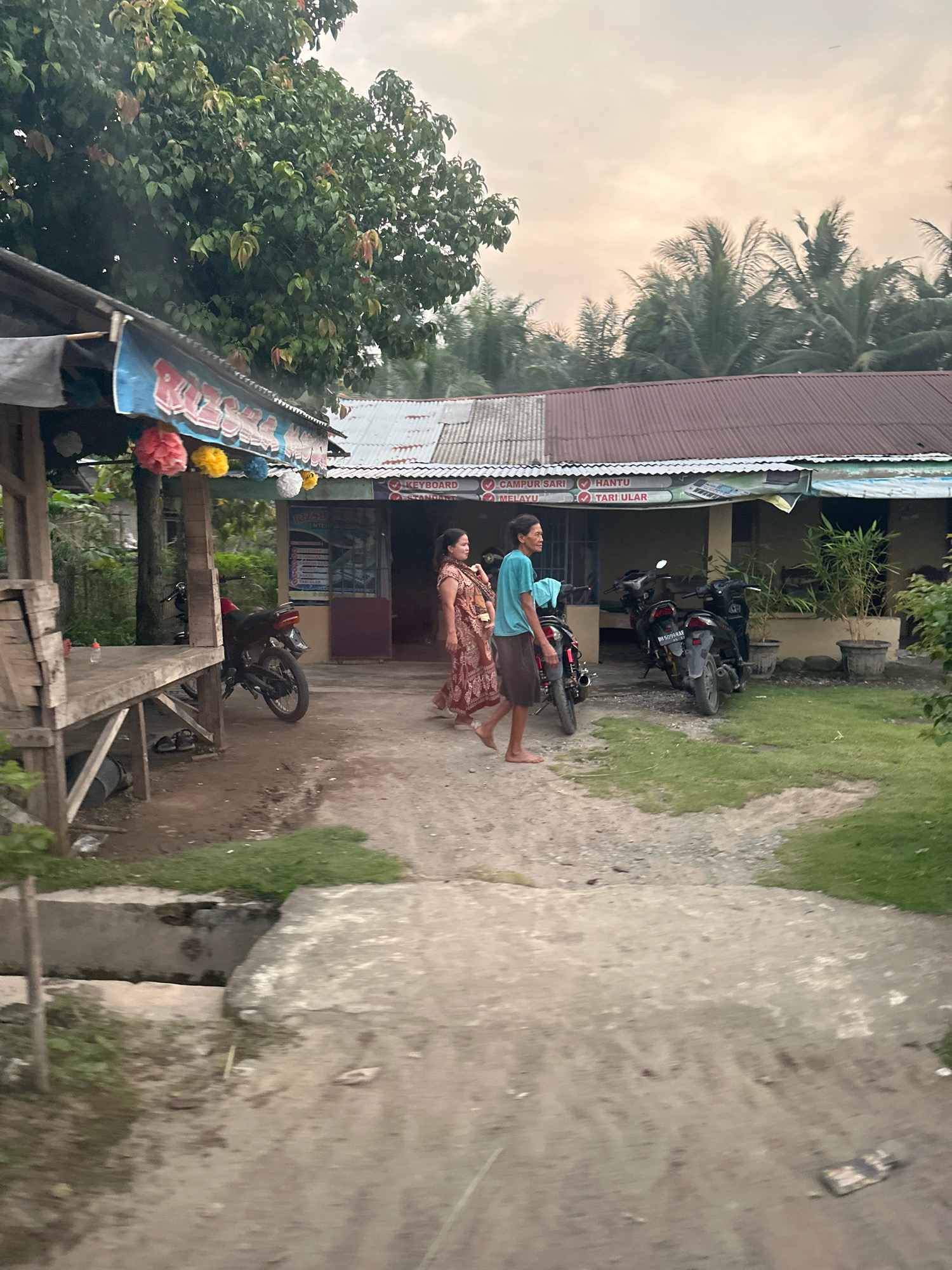 The image depicts a scene in front of a building in Tandem Hilir I, Indonesia. Two individuals, a woman wearing a patterned dress and a man in a teal shirt and brown shorts, are walking on a dirt path. To their left, a red motorcycle is parked near a wooden structure. The building behind them has a corrugated metal roof and appears to be a commercial establishment with signage that includes the words "CAMPUR SARI," "MELAYU," "HANTU," and "TARI ULAR." Several other motorcycles are parked in front of the building. The environment is somewhat overgrown with grass and trees, suggesting a rural or semi-rural setting. The sky is overcast, and the lighting indicates either early morning or late afternoon.