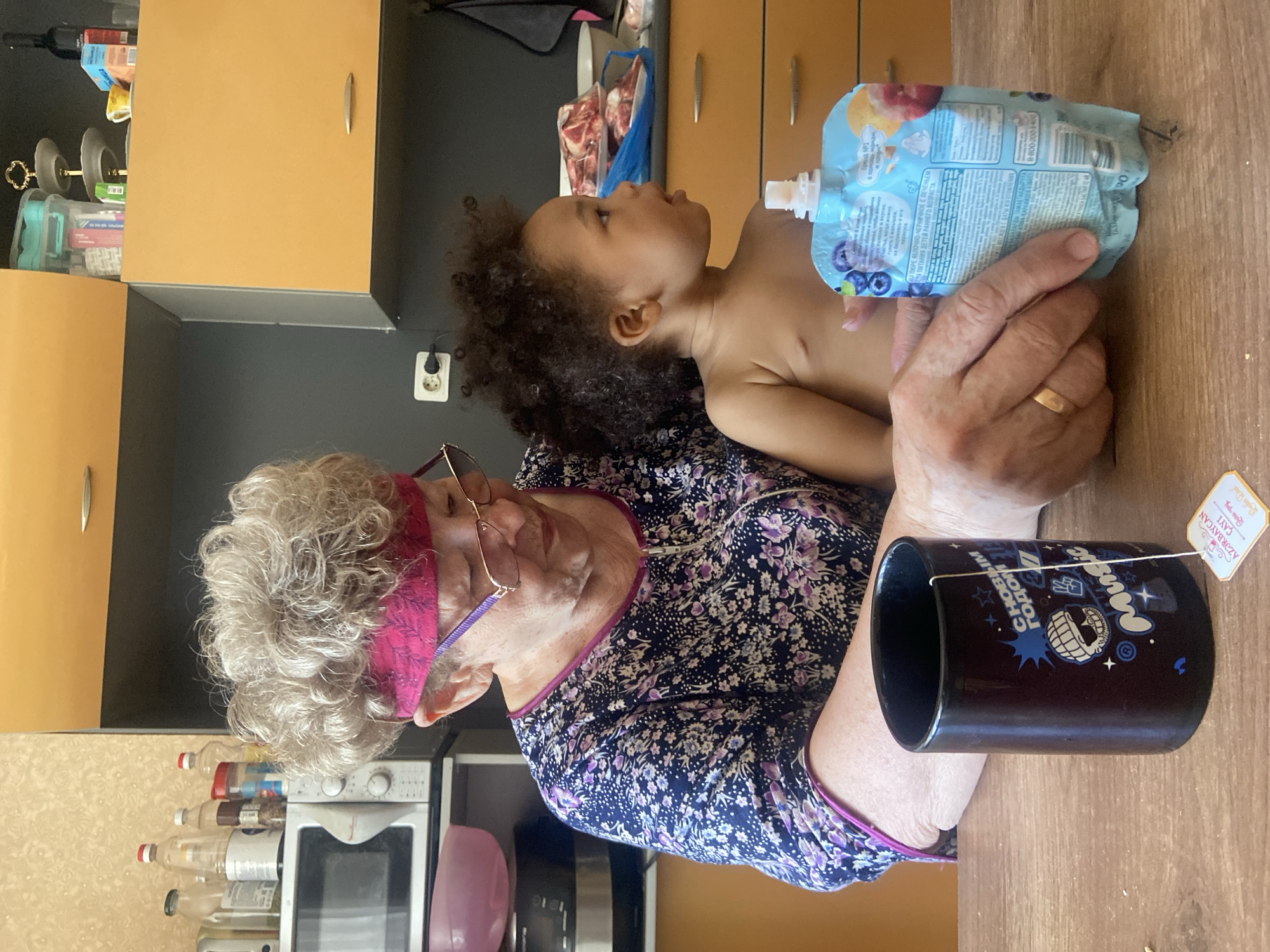The image shows an elderly woman indoors, seated at a wooden table, holding a small child on her lap. She is wearing a blue and purple floral top, glasses, and a pink headband. The child, who appears to be a toddler, is bare-chested and looking upwards. The woman is holding a blue baby food pouch toward the child. On the table, next to the woman, is a black mug with cartoon-like designs and the words "Music" and what appears to be "С НОВЫМ ГОДОМ" in Russian, along with a teabag tag written in Cyrillic script, mentioning “AZƏRBAYCAN ÇAYI.”
The background includes kitchen cabinets, a microwave, and various bottles and containers. Based on the product information visible in the image (the AZƏRBAYCAN ÇAYI tea) and the Russian script seen on the mug and baby food pouch, the location is in a country where Russian is commonly used, such as Russia. Given the specific Azeri tea mentioned, it is possible the scene is in Kazan, Russia, a multicultural city with a Tatar population.