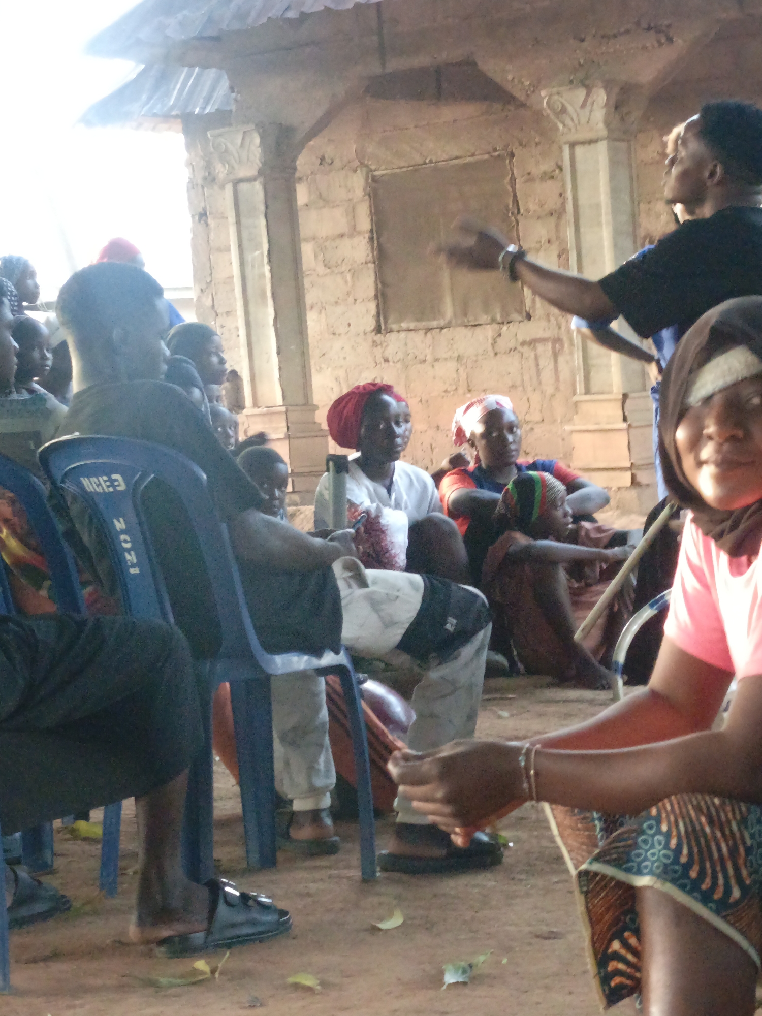 Akwanga, Nigeria: A gathering of people, including adults and children, are congregated outdoors. A man in a black shirt gestures with his arm raised. People are seated in chairs and on the ground. Building with brick walls, columns, and carvings in the background.