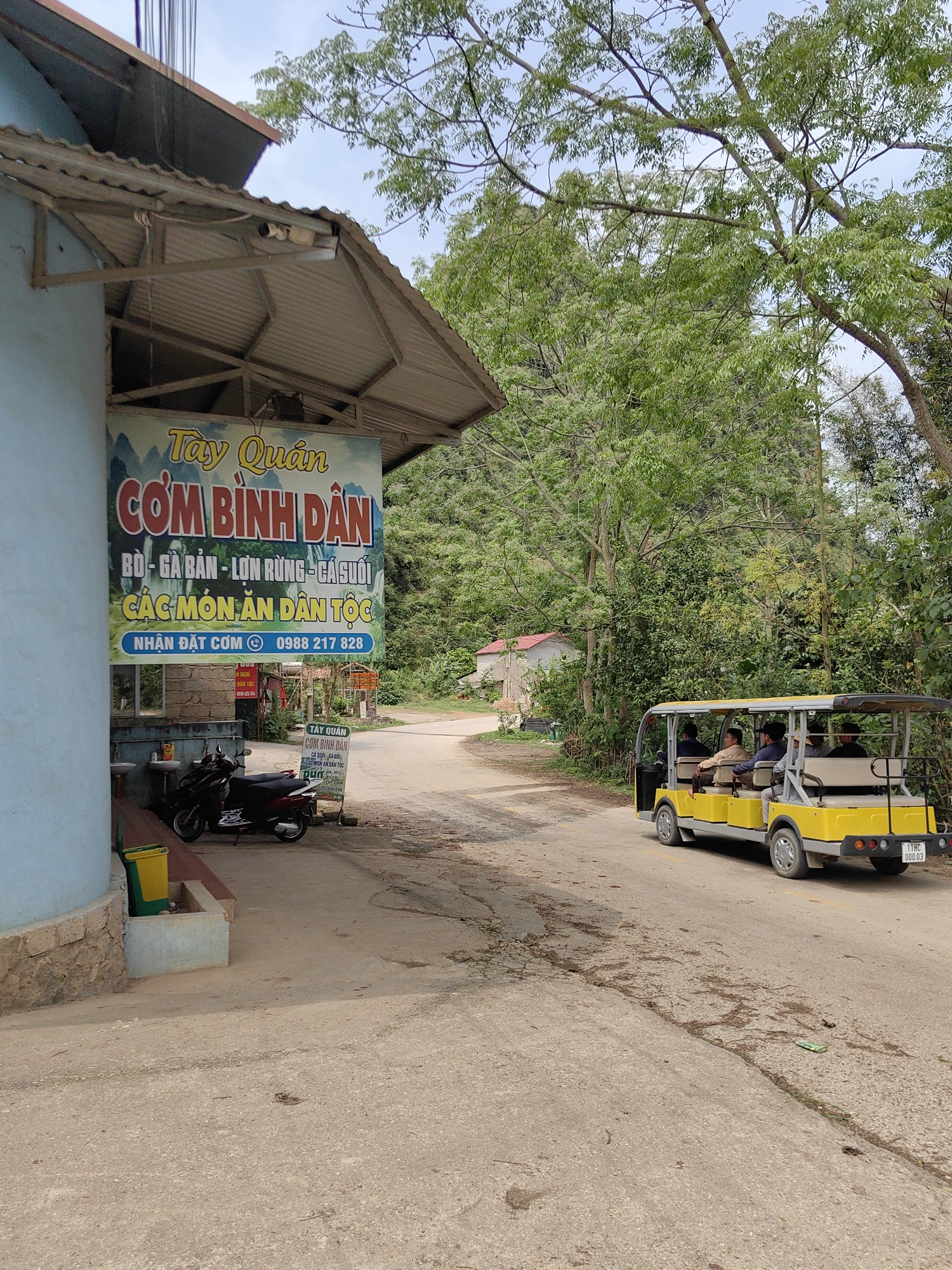 The image depicts a scene in Xã Đàm Thủy, Vietnam. A restaurant named "Tày Quán" advertises "Cơm Bình Dân" (affordable rice dishes), with a sign listing beef, chicken, wild boar, stream fish, and ethnic dishes. A phone number is displayed for ordering. A scooter is parked near the restaurant entrance. A yellow transport vehicle carrying passengers is on the road. The environment consists of trees, a building in the background, and a paved road.