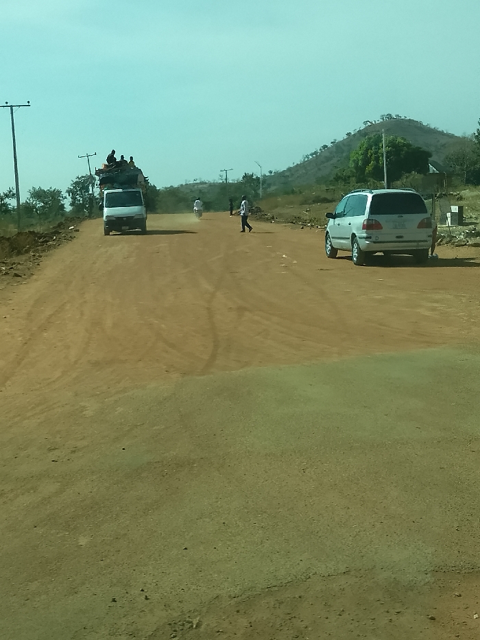 The image depicts an unpaved, reddish-brown dirt road in Jibam, Nigeria, under a clear, light-blue sky. Tire tracks are visible across the dusty road surface. A white delivery van, heavily loaded with cargo and multiple passengers seated atop its roof, travels away from the viewer on the left side of the road, generating a dust trail. Further down the road, a motorcycle with a single rider also moves away from the viewer, creating a smaller dust cloud. A pedestrian wearing a light-colored shirt is observed walking from right to left across the mid-ground of the road. On the right, a silver minivan is stationary, oriented away from the camera. The background features a gentle hill with sparse vegetation and multiple utility poles supporting overhead lines. Additional indistinct human figures are present further down the road.