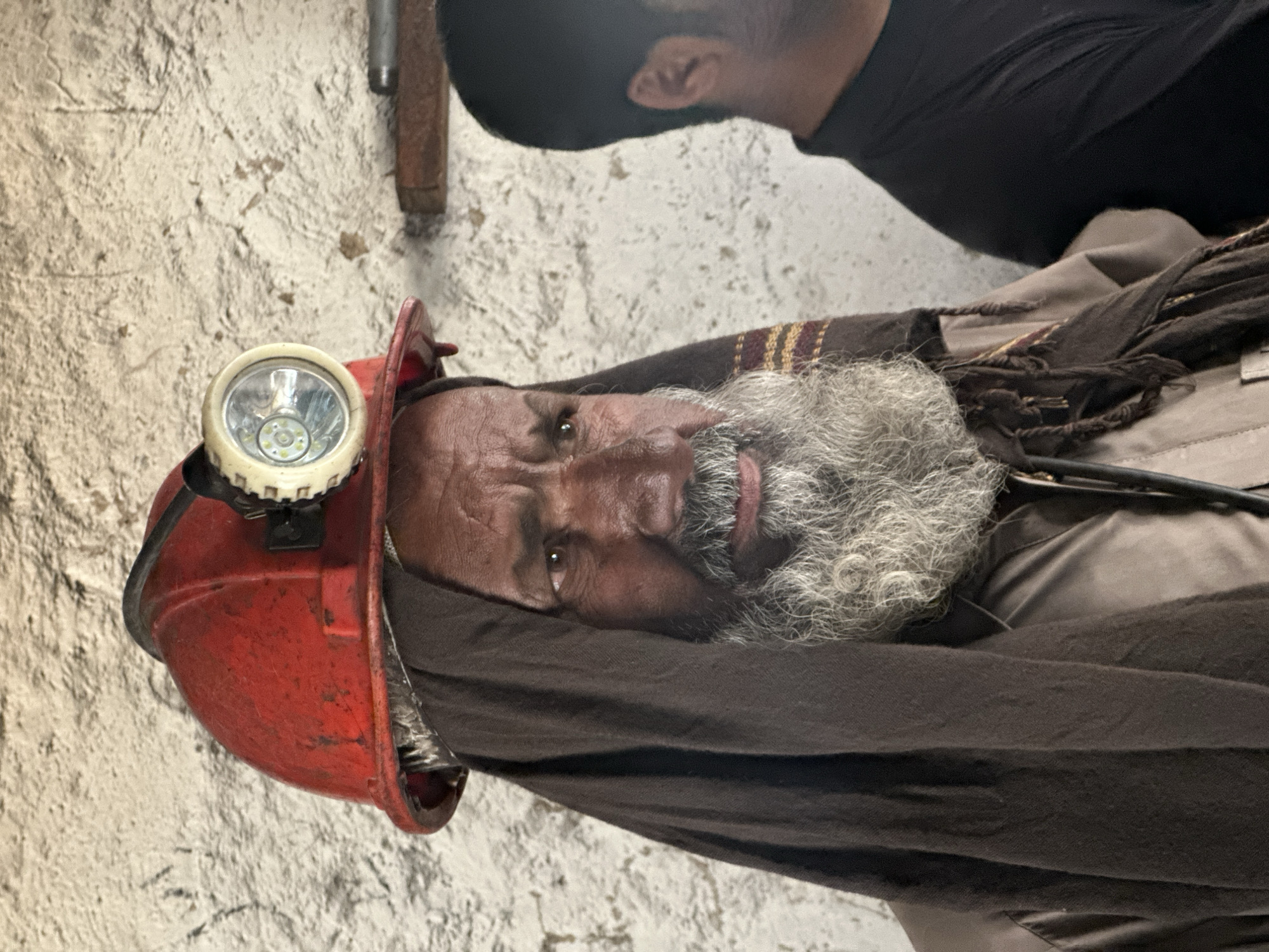 A close-up, medium shot captures a man wearing a red miner's helmet with a headlamp. The man has a greying beard and is looking to the right of the frame. He is also wearing a dark brown headscarf draped over his head and shoulders, which covers the lower part of his face and his neck. He is wearing a brown shirt. In the background, to the right of the man, is the shoulder and head of another person wearing a black shirt. The background wall appears to be made of rough, textured stone or plaster. The overall impression is a candid portrait of a miner, likely in Quetta, Pakistan, given the context.
