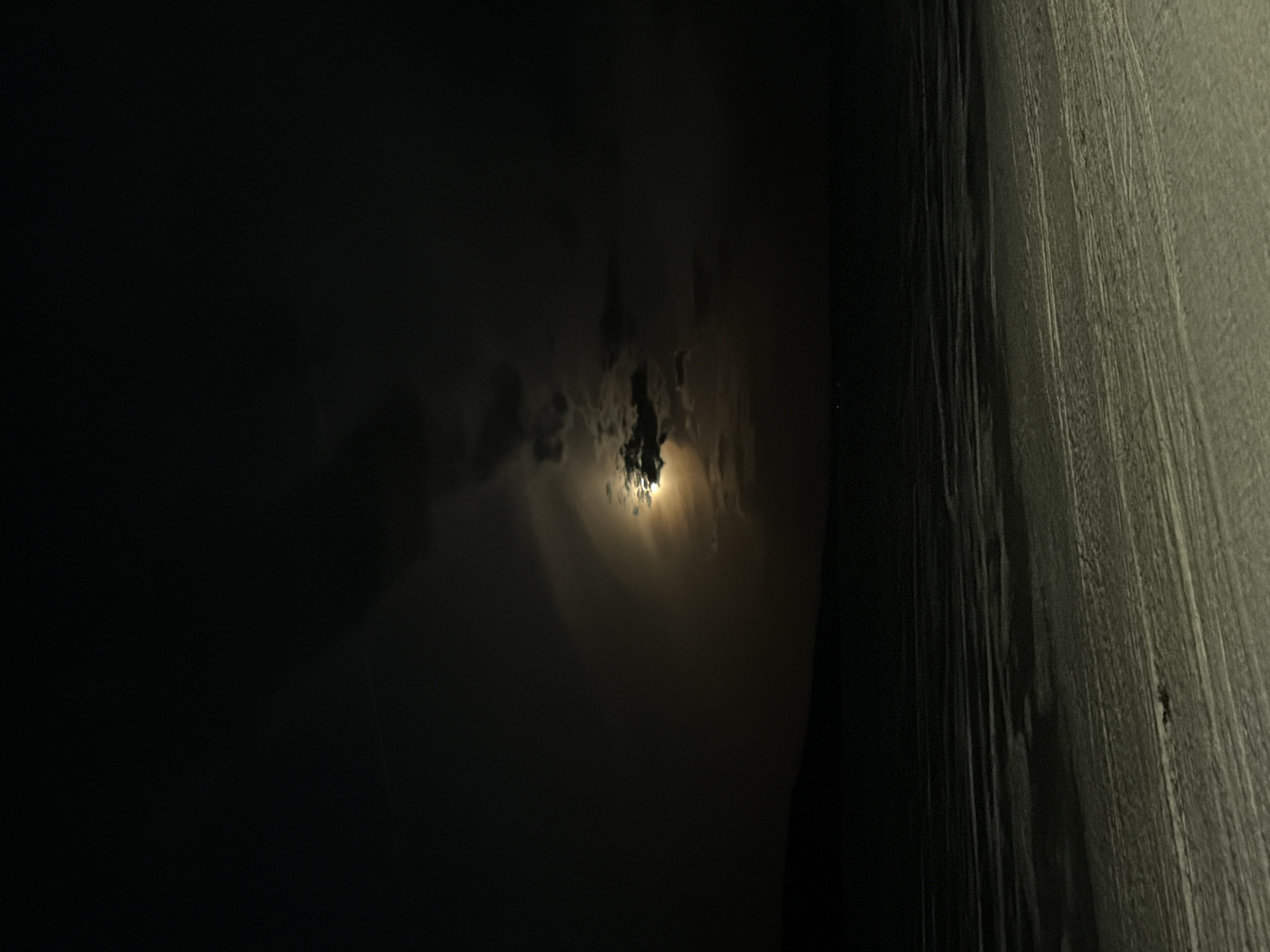 The image depicts a nighttime scene in Chaikovsky, Russia. The moon is partially obscured by clouds, casting a soft, diffused light. To the right, a tall, textured surface is visible, possibly a building or wall. The rest of the scene is shrouded in darkness. No people or specific objects are identifiable.