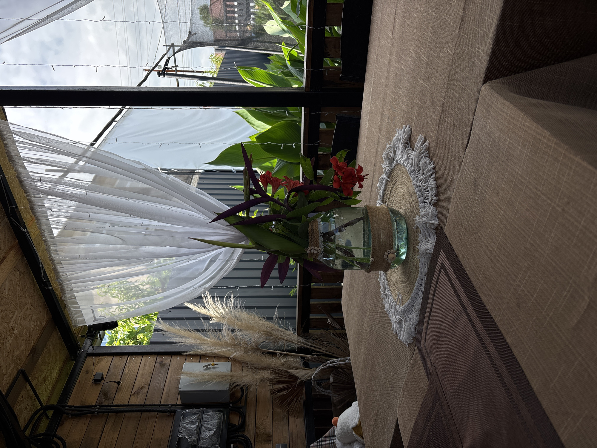 The image depicts a table set for a meal on a covered patio in Gudauta, Abkhazia. The table is covered with a brown tablecloth, and a vase containing red flowers and greenery sits on a straw placemat in the center. In the background, sheer white curtains hang from the ceiling, revealing lush green foliage and a cloudy sky beyond. Wooden walls and electric cables are visible on the left, suggesting an outdoor dining area. The overall scene conveys a sense of relaxed outdoor dining.