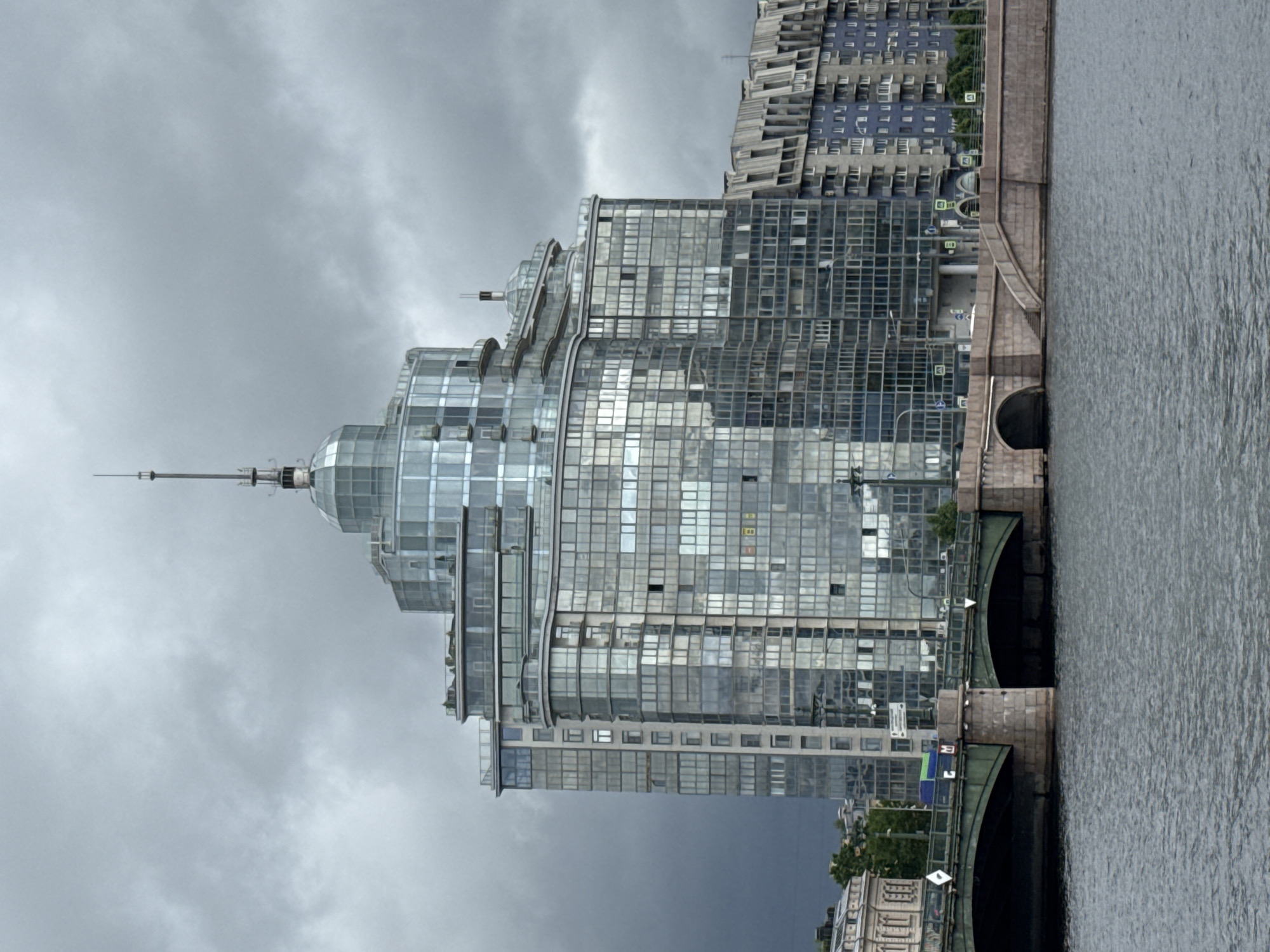 The image shows a modern glass-clad building in Saint Petersburg, Russia, likely situated along a riverfront. The building has a distinctive tiered structure with a central tower topped with a spire. The architecture appears to be contemporary, with an emphasis on glass and steel. Adjacent to the building is a bridge spanning a body of water, with a visible archway below. The sky is overcast and cloudy, indicating potentially inclement weather. Other buildings can be seen in the background.