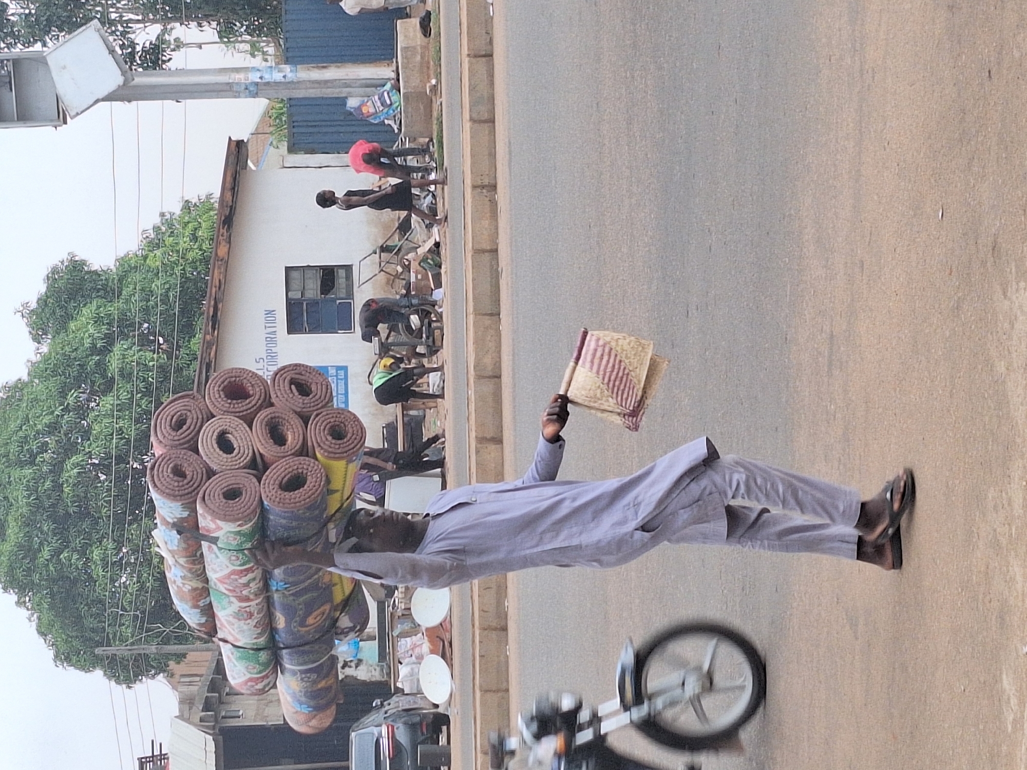In Sabon Tasha, Nigeria, a man carries a stack of rolled mats on his head while walking along a paved road. He holds a hand fan in his other hand. He is wearing a light-colored, long-sleeved shirt, pants, and sandals. In the background, there are people, buildings, and a motorcycle.