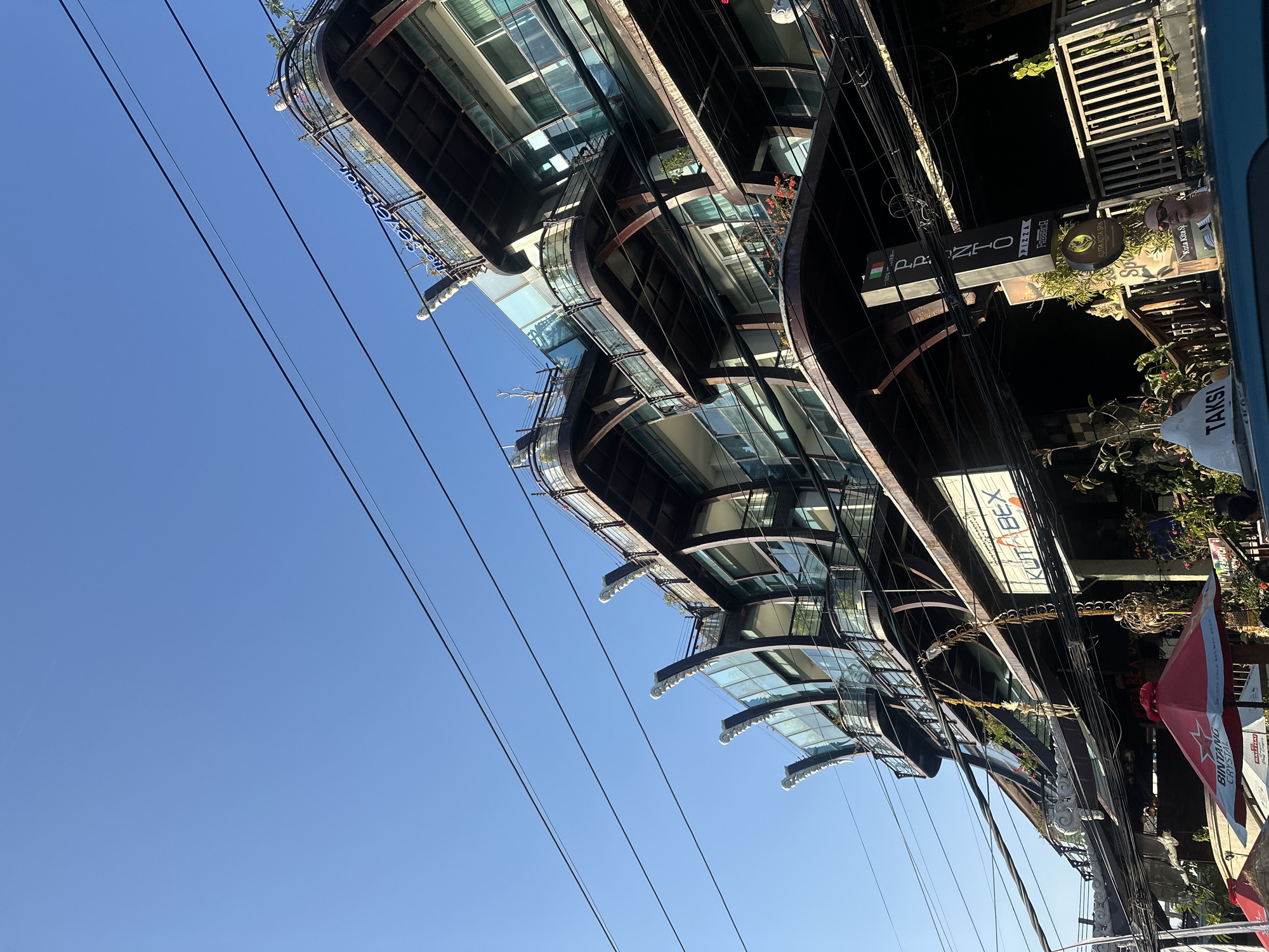 The image shows a multi-story building in Kuta, Indonesia, with a distinctive curved architectural design. The building has several balconies with glass railings, and the facade is a mix of glass and dark-colored materials, possibly metal. There are numerous power lines crisscrossing in front of the building. Several signs are visible, including one that reads "KUTABEX". On the street level, there are umbrellas, plants, and a portion of a vehicle with a "TAKSI" sign on its roof. The sky is clear and blue. The building appears to be a commercial establishment, possibly with shops or restaurants on the lower floors and apartments or hotel rooms on the upper floors. The overall scene suggests a busy urban environment in Kuta.