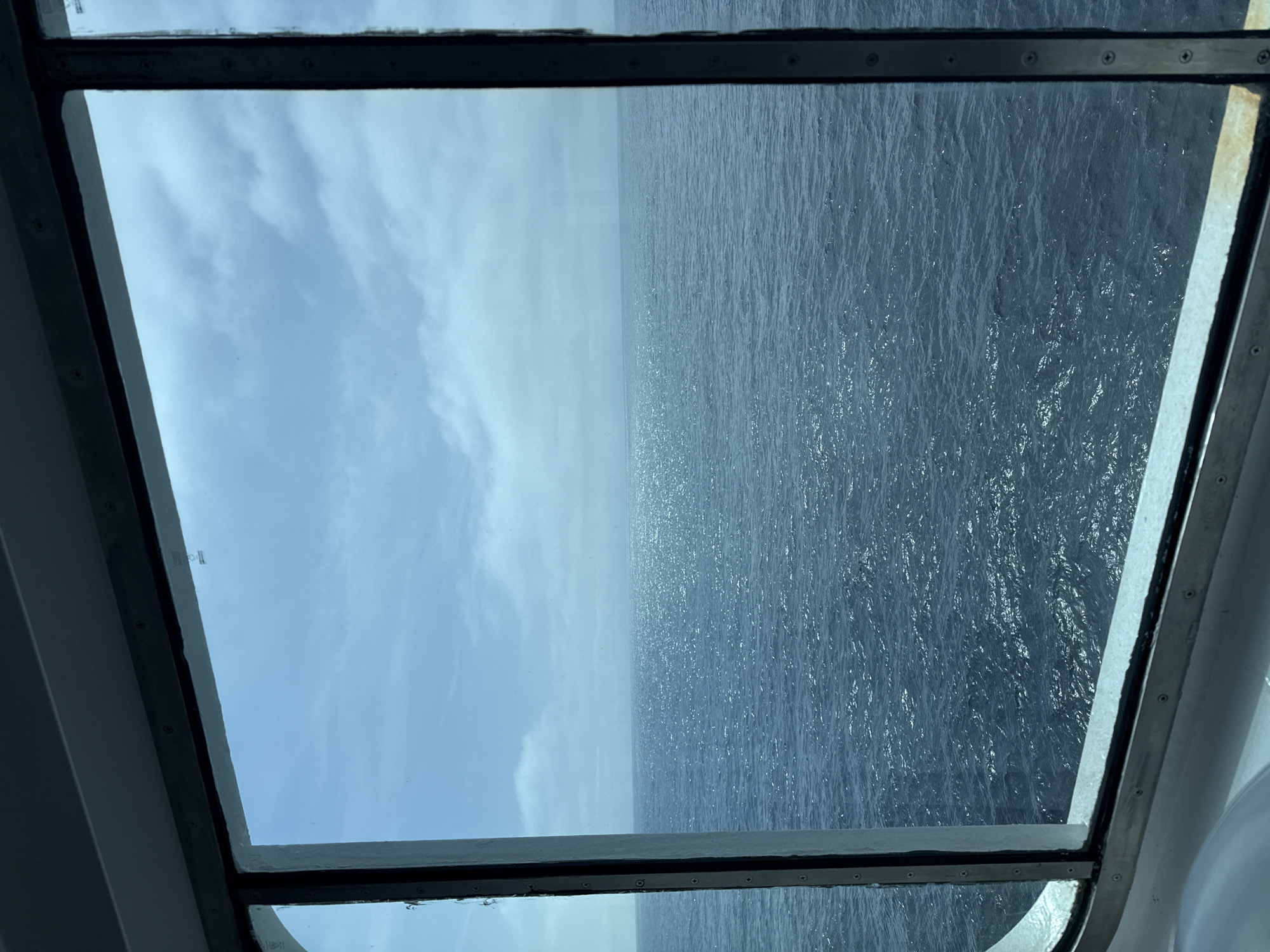The image presents a view through a window, showing the sky and the ocean. The sky, on the left side of the frame, is a pale blue with scattered clouds. The ocean, on the right side, appears dark blue with subtle ripples and glimmers of light reflecting on the surface. The window frame is visible around the edges of the image, suggesting the viewpoint is from inside a structure, likely a ship or building near the water. The location is unknown.