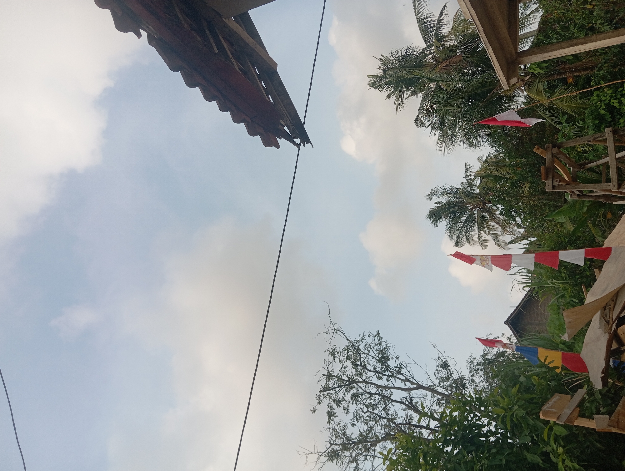 The image depicts an outdoor scene with a cloudy sky. In the foreground, there are several flags with red and white stripes, as well as a blue and yellow flag, positioned near some wooden structures and greenery. A house with a dark roof is visible in the background, partially obscured by trees. To the right, a portion of a building with a tiled roof is visible, showing some damage. A power line runs horizontally across the upper part of the frame. The environment appears to be a rural or semi-rural area in Dente Teladas, Indonesia.