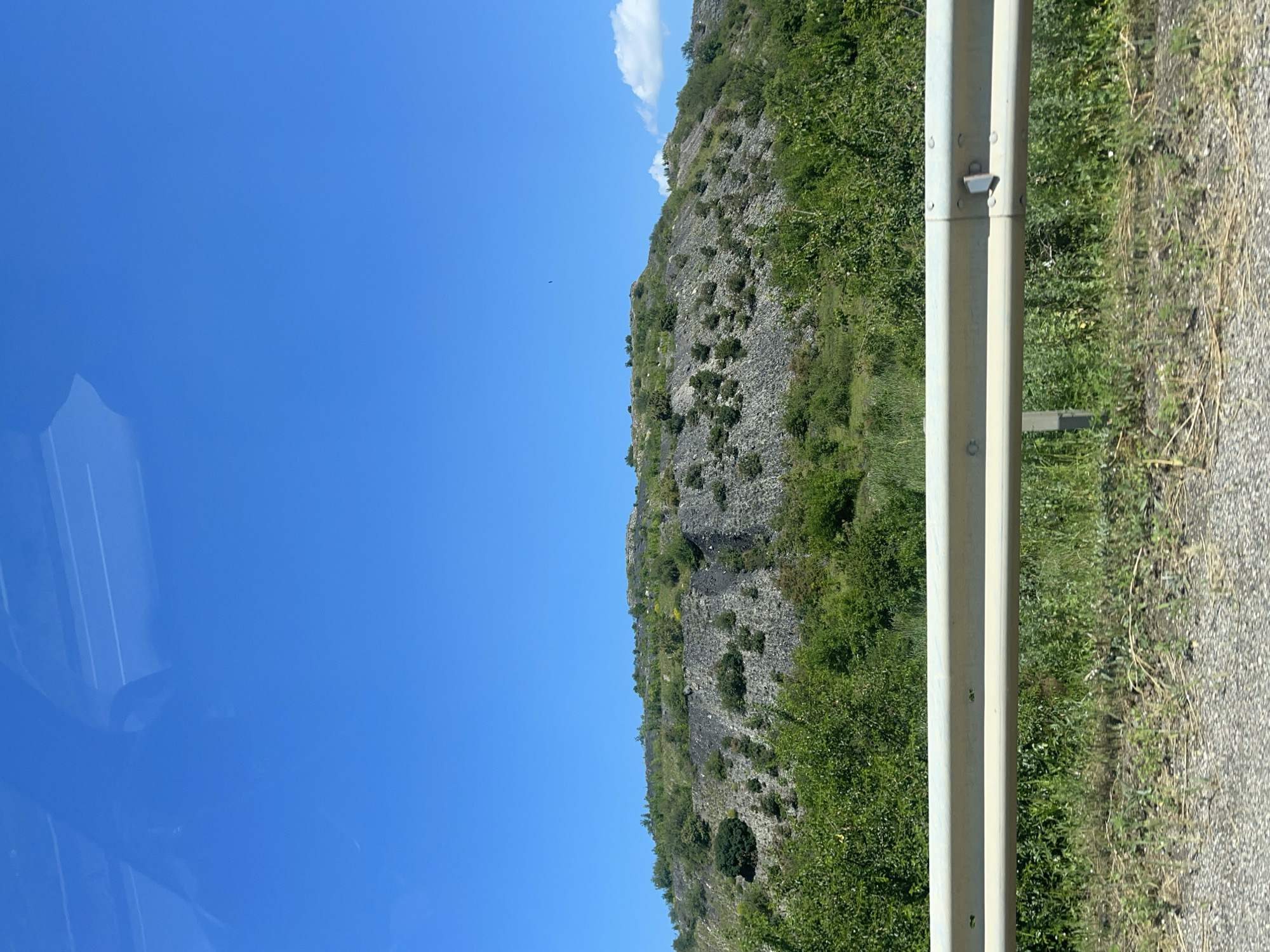 The image shows a barren, rocky hillside dotted with sparse green vegetation. A clear blue sky stretches across the top of the frame, with a few wispy clouds on the right side. In the foreground, a guardrail runs horizontally across the image, separating the viewer from the hillside. The ground in front of the guardrail is covered in gravel and dry grass. The overall impression is one of a rugged, natural landscape under a bright, sunny day. There are no people or specific landmarks visible in the image that would indicate a specific location in Загорский, Russia.