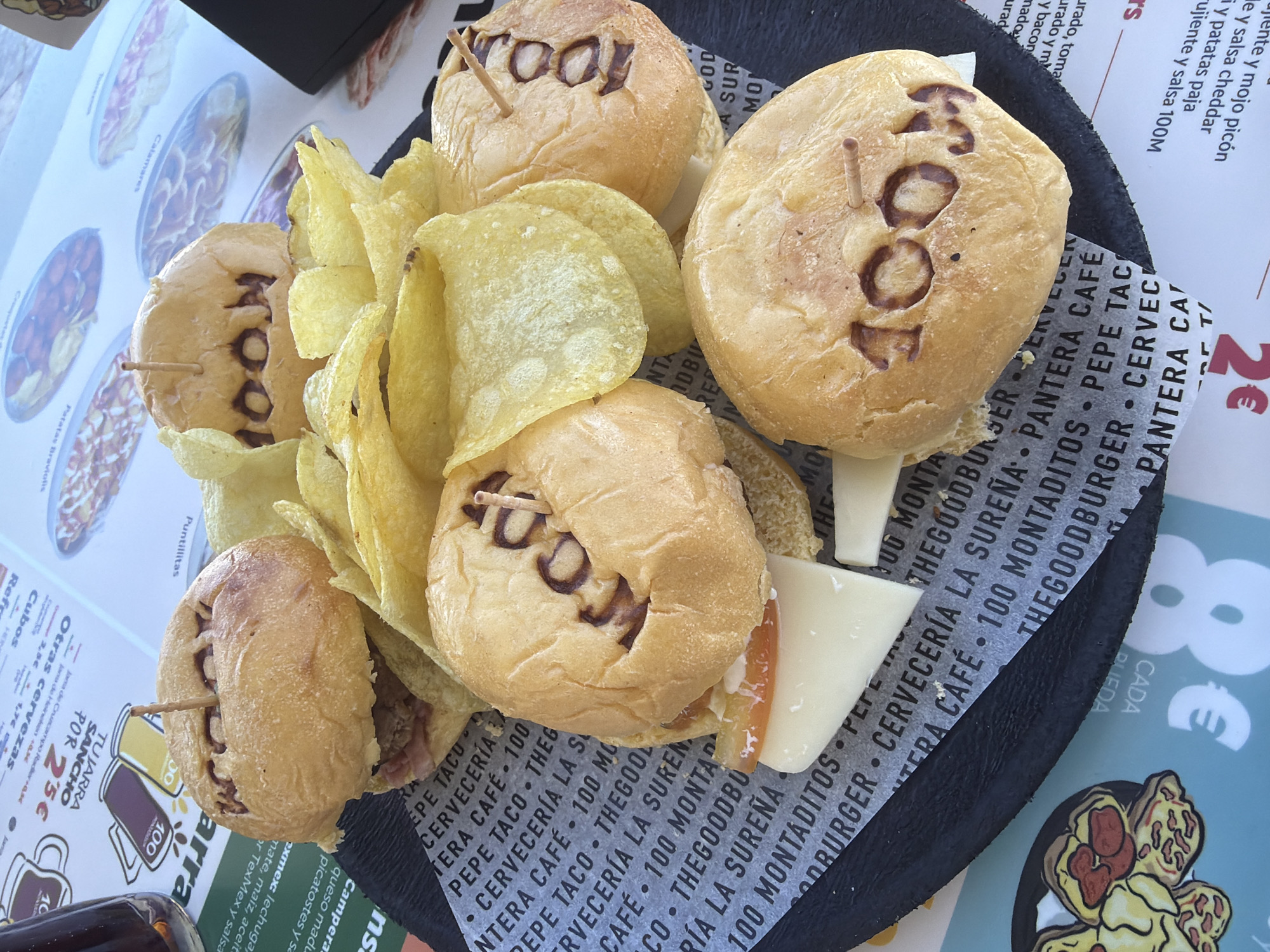 The image presents a platter of five mini-burgers labeled "100," accompanied by potato chips and cheese slices. The burgers are secured with wooden toothpicks. The platter sits on a printed paper with text in Spanish that includes the phrase "CERVECERIA LA SURENA", indicating the name of a restaurant. The paper also features logos and the words "PEPE TACO" and "THE GOOD BURGER." The table beneath the platter is covered with a printed menu, which includes dish names like "Calamares" and "Patatas Bravas". The menu is priced in euros. Given the restaurant names and the menu items, it is highly likely this image was taken in Alicante, Spain.