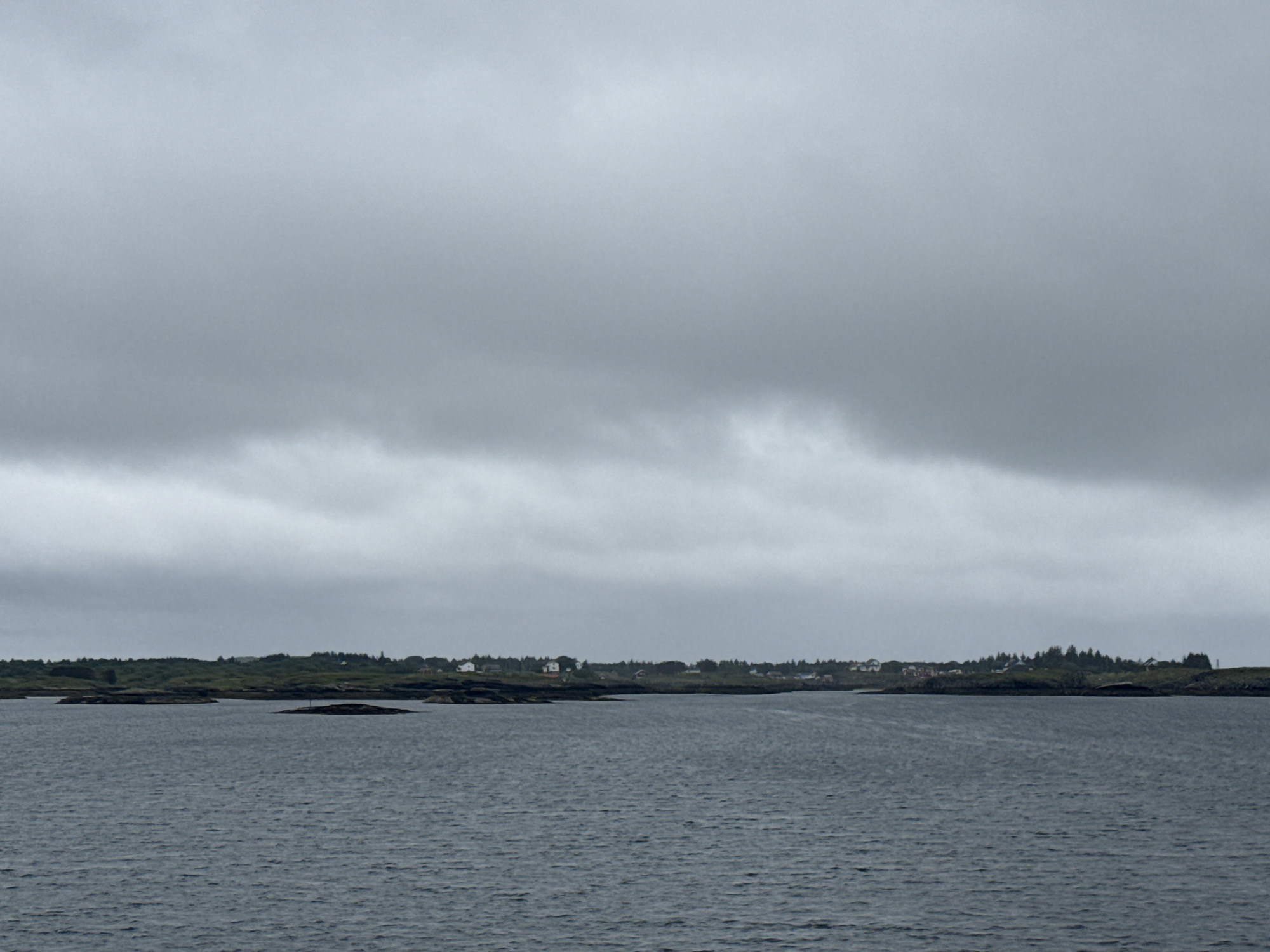 The image depicts a calm, dark body of water with a slightly choppy surface, reflecting the overcast sky. In the distance, a low-lying island or coastline is visible, characterized by dark, rocky outcrops interspersed with green vegetation. A scattering of small houses, some white and others with red roofs, are nestled among the trees on the higher ground of the island. The sky is a uniform, pale gray, heavily clouded, suggesting a cool, possibly rainy day. The overall atmosphere is one of muted natural beauty and quiet habitation. This scene is consistent with the coastal landscape of Sleneset, Norway, known for its scattered islands and maritime environment.