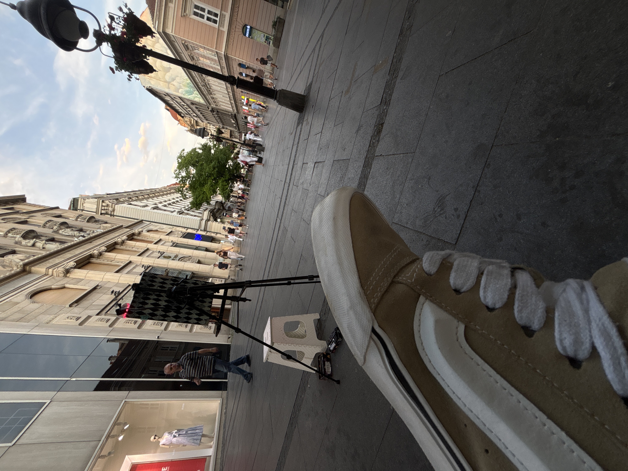 This is a first-person perspective, low-angle shot of a street scene in Belgrade, Serbia. In the foreground, a tan canvas shoe with white laces is prominent, filling the bottom left portion of the frame. The shoe is partially obscuring the cobblestone pavement, which is made of dark grey rectangular stones.
To the left of the shoe, a white plastic stool is positioned on the pavement. Next to the stool, on the ground, are several items including what appears to be a small box labeled "TIPS" and some batteries. Behind these objects, a tripod stands, supporting a black and patterned screen or board.
Further back in the mid-ground, a man in a striped t-shirt and jeans walks away from the camera. To his left is a building with a large glass storefront, displaying a mannequin in a light blue dress. The building facade is a light grey material, reflecting the sky and surrounding structures.
The background depicts a bustling city street with pedestrians walking in both directions. The architecture features ornate, multi-story buildings with classical elements. A lamppost with hanging flower baskets is visible on the right side of the street. The sky above is a pale blue with scattered white clouds, suggesting it is late afternoon or early evening. The overall impression is of a lively urban environment.