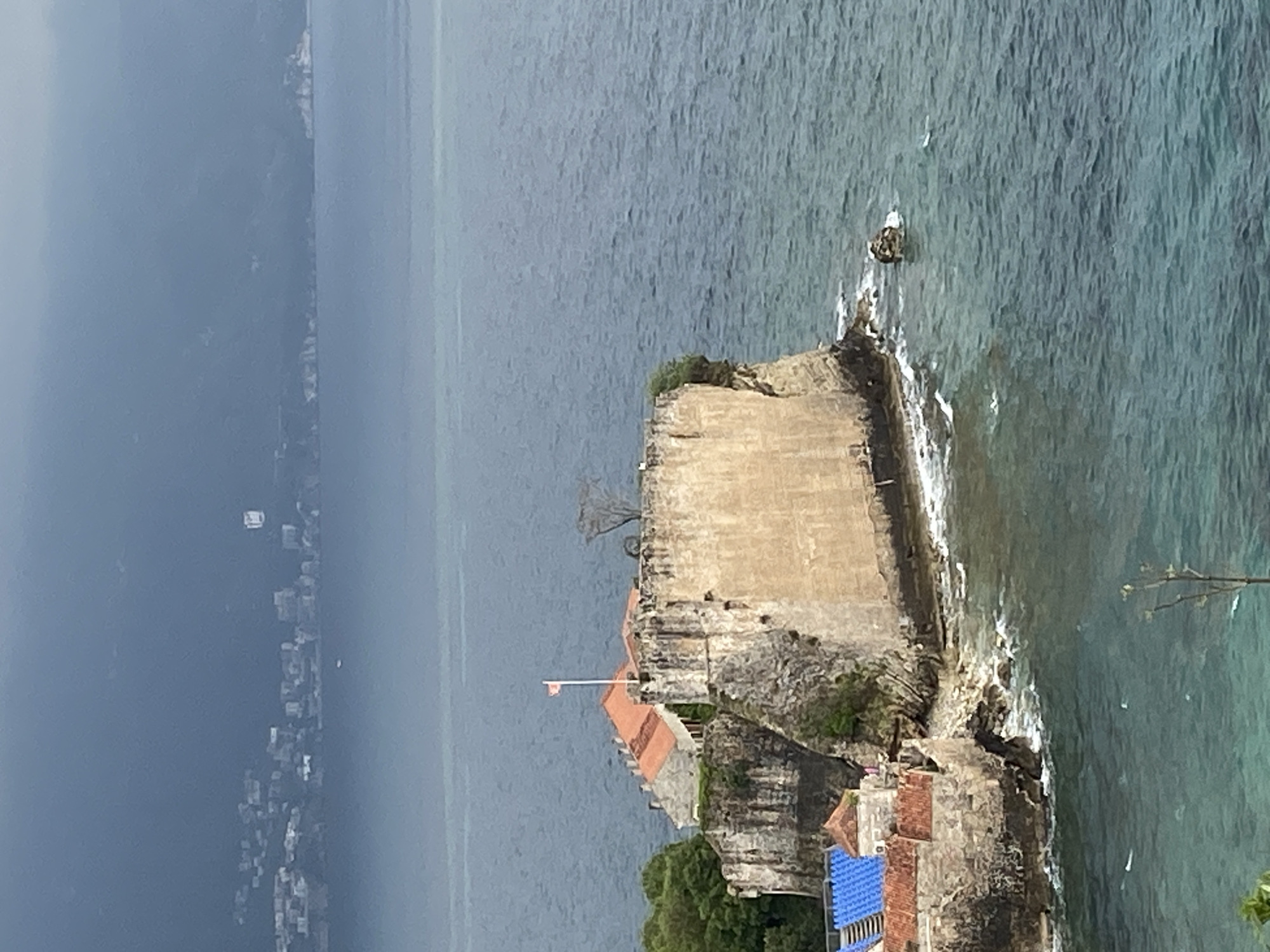 The image shows a high-angle, slightly rotated view of a coastal area near Budva, Montenegro. A large stone structure, possibly a fortress or historical building, dominates the foreground and extends into the sea. The structure is tan and gray, with signs of age and weathering. Parts of the structure have red tile roofs. A white flag is on top of the structure. The sea is visible on three sides of the structure, showing varying shades of blue, indicating different depths and conditions.
In the distance, the coastline curves, with a cluster of buildings and a hillside visible under an overcast sky. The water appears somewhat turbulent around the base of the structure, with small waves crashing against the stone. There is also evidence of foliage and plant growth on top of and around the base of the stone structure. A small boat is located in the water to the right of the structure.
