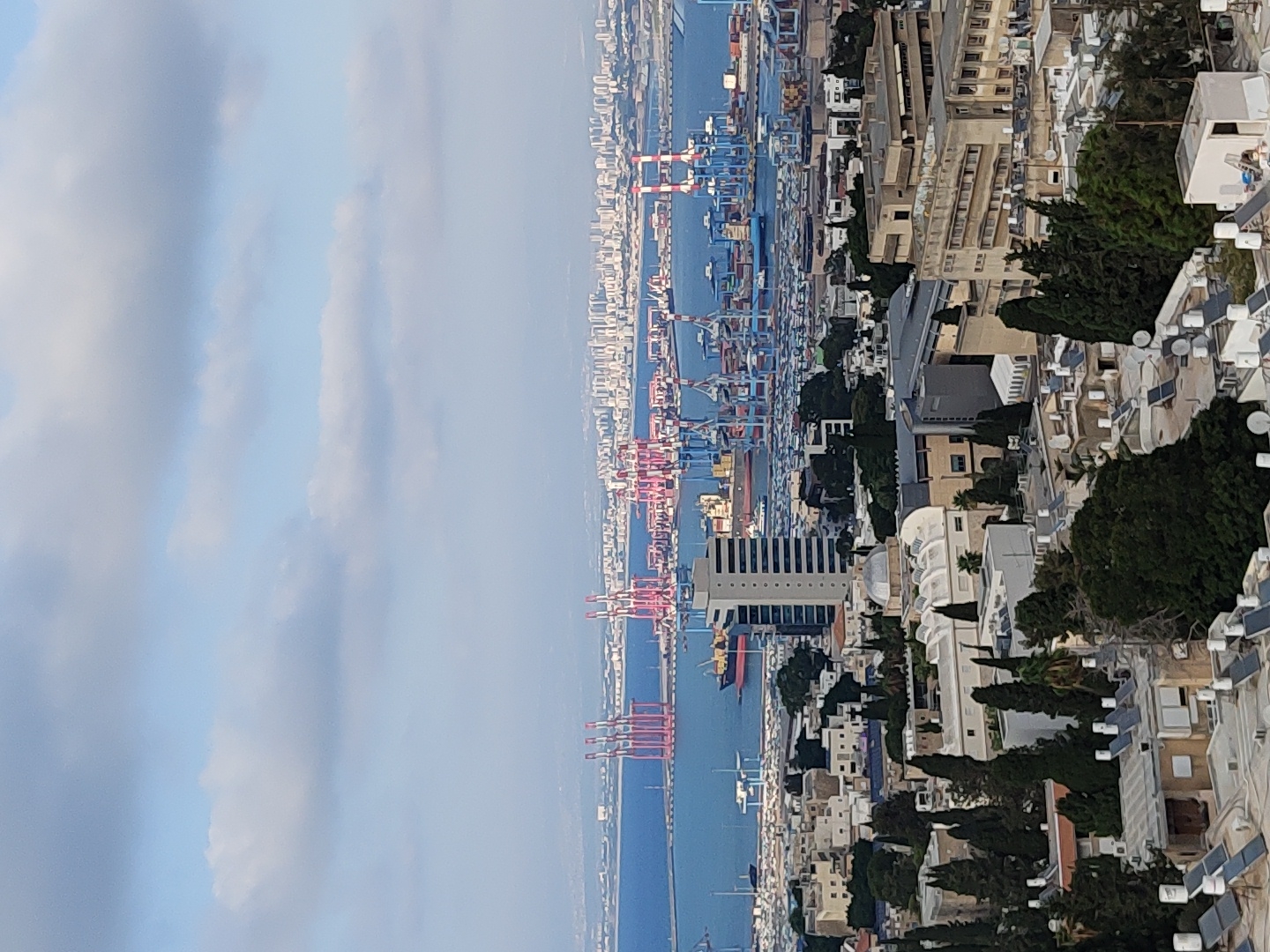 This is an aerial, medium-distance shot of the city of Haifa, Israel. The image captures a panoramic view of the city, a large harbor, and the sea, all under a partly cloudy sky. In the foreground, residential buildings with flat rooftops are visible, many equipped with solar panels and satellite dishes. Among the buildings, some appear older with lighter colored stone facades, while others are modern constructions. Lush green trees are interspersed throughout the urban landscape.
Further in the mid-ground, the distinct architecture of a domed building, likely a public or religious structure, stands out. Adjacent to it are more institutional-looking buildings with multiple stories and large windows, suggesting administrative or educational facilities. A prominent tall, rectangular building with a light-colored facade also features in this area.
The background is dominated by the expansive Haifa Bay port. Numerous large, red and blue container cranes are lined up along the docks, indicating active cargo operations. Several cargo ships and container vessels are moored in the bay. Beyond the port, a dense urban skyline stretches along the coastline, representing the city's commercial and residential areas. The sea itself is a deep blue, merging with the horizon where a hazy, distant landmass is visible. The overall scene conveys a sense of a bustling port city with a mix of historical and modern architectural elements.