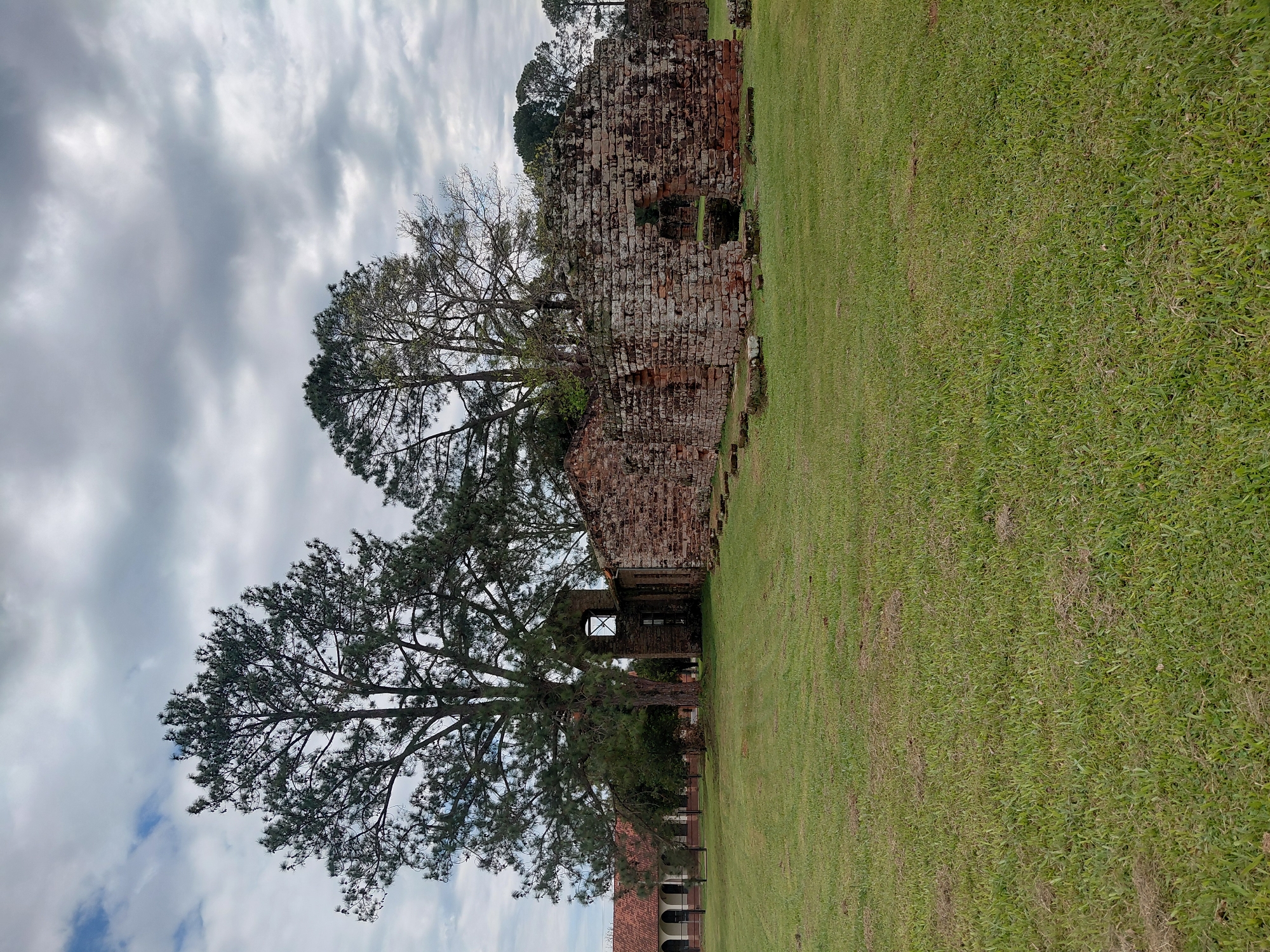 The image captures a historical ruin in Trinidad, Paraguay, under a cloudy sky. In the foreground, a vast expanse of green grass stretches across the scene. To the right, the weathered stone walls of a ruin are visible, characterized by their rough-hewn stones and mossy surfaces. Several arched openings punctuate the walls, hinting at its former structure. Behind the ruin, a large tree with dense foliage stands prominently, its branches reaching towards the sky. In the background, a long, red-tiled building with arched windows and columns suggests a more preserved historical structure, possibly part of the same complex. The overall environment is serene and historical, evoking a sense of the past.