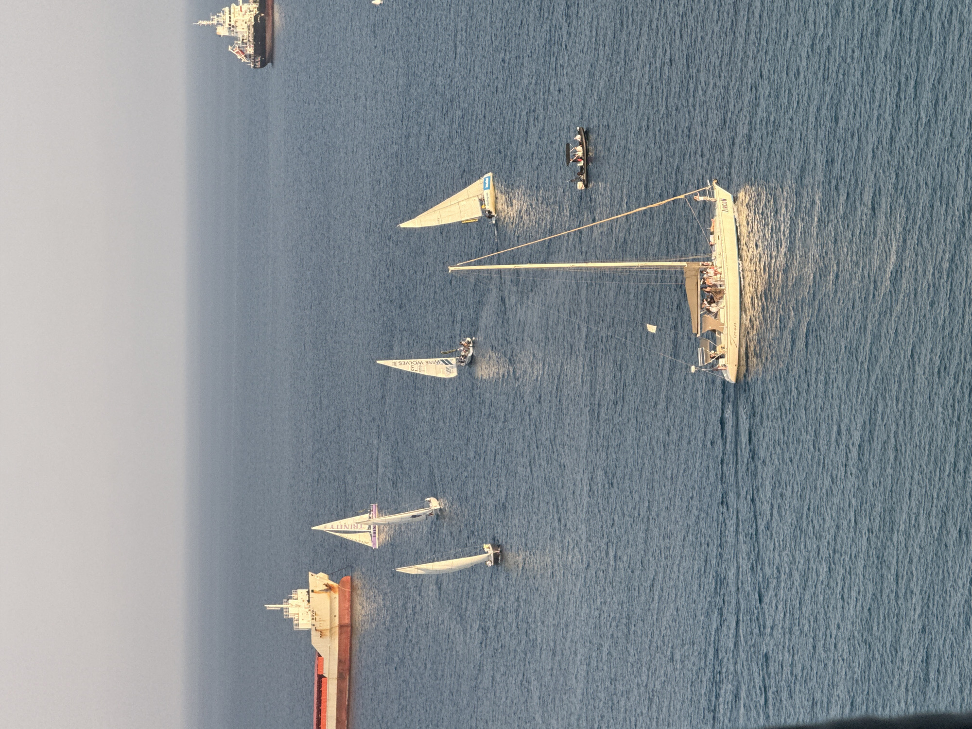 This is an aerial, eye-level shot of the sea off the coast of Limassol, Cyprus, during the day. The water is a deep blue, with some gentle waves. In the background, a large cargo ship is visible on the left, and a smaller white ship, possibly a pilot boat or tug, is on the right. In the middle ground, several sailboats are racing. One sailboat, with the name "ZIRCON" visible on its hull, is in the foreground, with many people on deck. A small motorboat is also present, carrying at least two individuals. The sky is a pale, hazy blue. The image captures a serene maritime scene, likely during a regatta or sailing event.