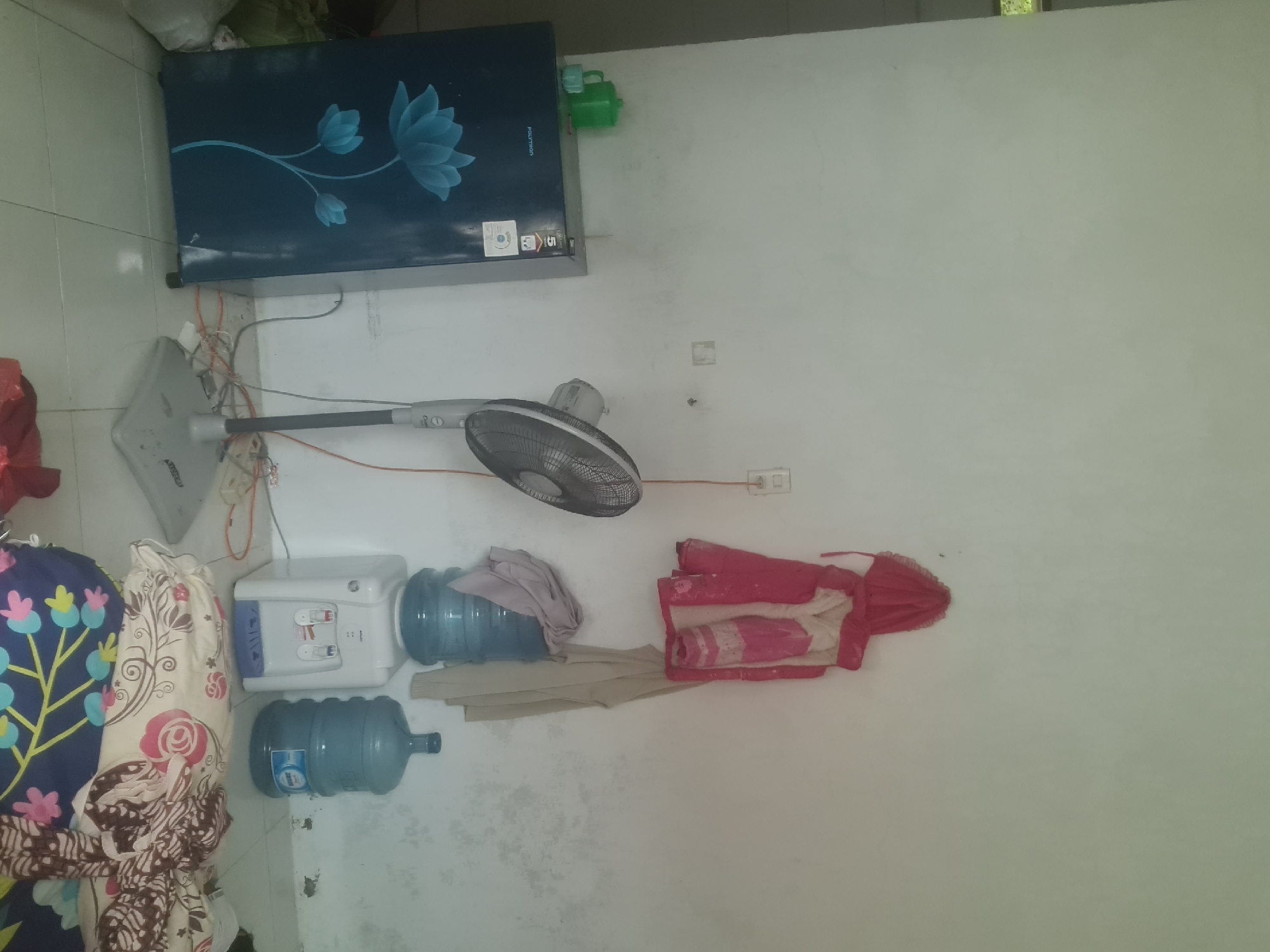 The image depicts an indoor scene within a room, possibly a domestic dwelling or a small shop, located in Lhokseumawe, Indonesia. The room is sparsely furnished and appears somewhat cluttered. A prominent dark blue refrigerator with a floral design is positioned on the left side of the frame. To the right of the refrigerator, a standing fan is visible. Above the fan, two water cooler bottles are stacked on top of a white water dispenser. Hanging on the wall above the water dispenser are two pieces of clothing, one a pink hooded jacket and the other a beige garment. The walls are painted white and show signs of wear and discoloration. A power outlet and electrical cords are visible on the wall. In the foreground, a stack of folded fabric, likely bedding or blankets with floral patterns, rests on the tiled floor. The overall impression is one of a functional, lived-in space.