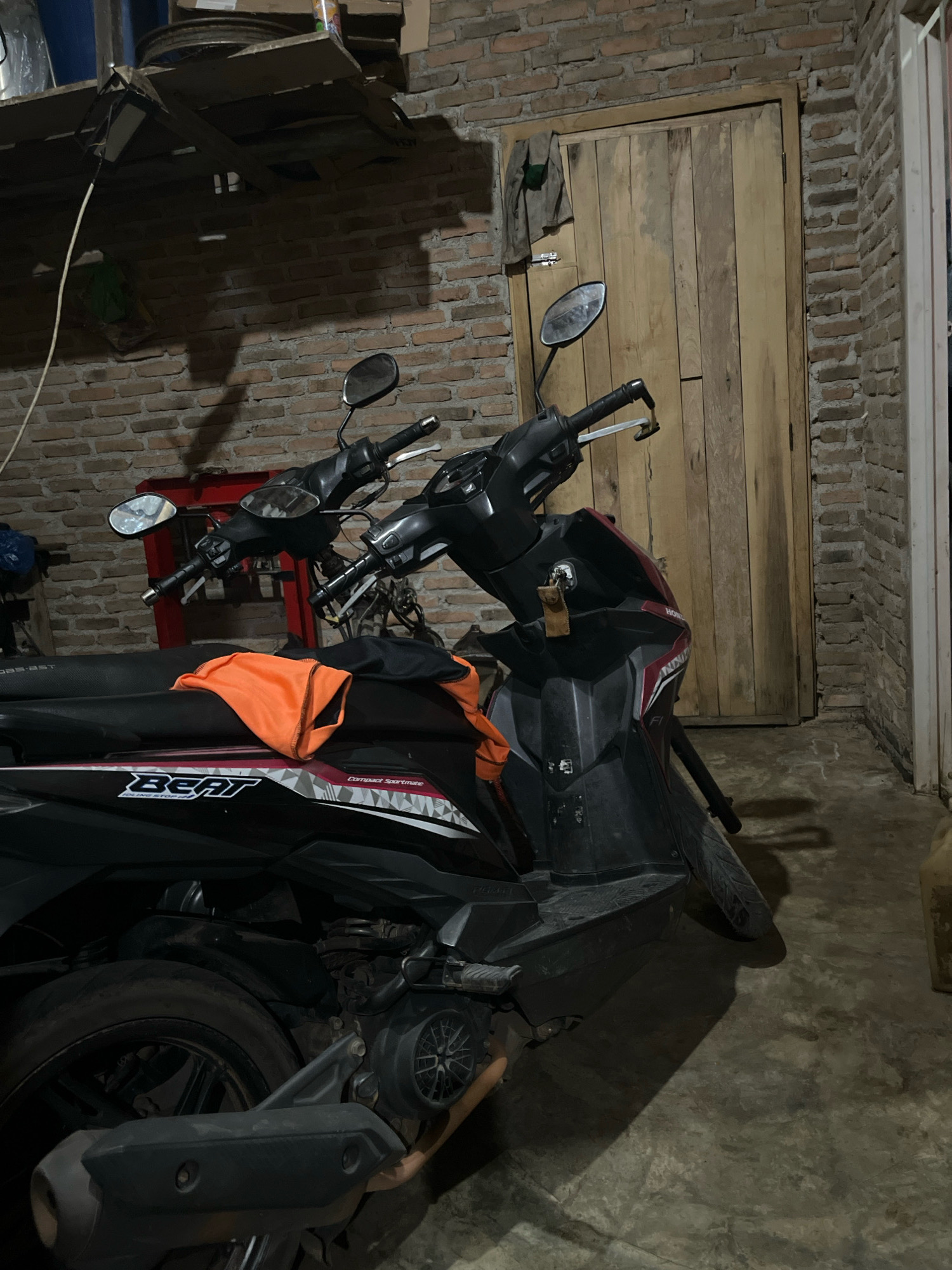 The image depicts an indoor scene, likely a garage or workshop, with a red brick wall and a wooden door as the backdrop. Two black and red scooters are parked in the foreground, one partially obscured by the other. An orange cloth is draped over the seat of the closer scooter, which has "BEAT" written on its side. The floor is concrete, and the lighting suggests it's nighttime or a dimly lit area. In the upper left corner, a shelf holds various items, including what appears to be a bicycle wheel rim. A shadow of a bicycle is cast on the wall. The overall impression is one of a functional, somewhat cluttered space. There are no people visible in the image.