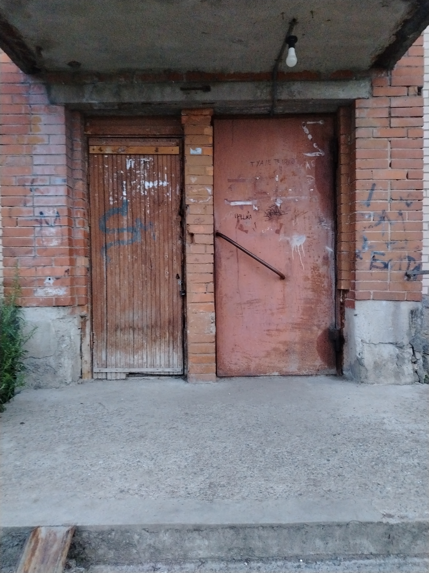 The image shows the entrance to a building with two doors. The left door is made of vertical wooden slats and is painted brown. It has some graffiti on it. The right door is made of metal and is painted a reddish-brown color. It also has graffiti and a metal handle attached diagonally across it. Both doors are weathered and appear old. The entrance is made of concrete and is flanked by brick walls. There is a light bulb hanging from the ceiling above the entrance. The scene is outdoors, possibly in an alleyway or a less-maintained part of a city. The overall impression is one of neglect and age. Vyborg, Russia is a port city in Leningrad Oblast, known for its medieval castle and historic architecture.