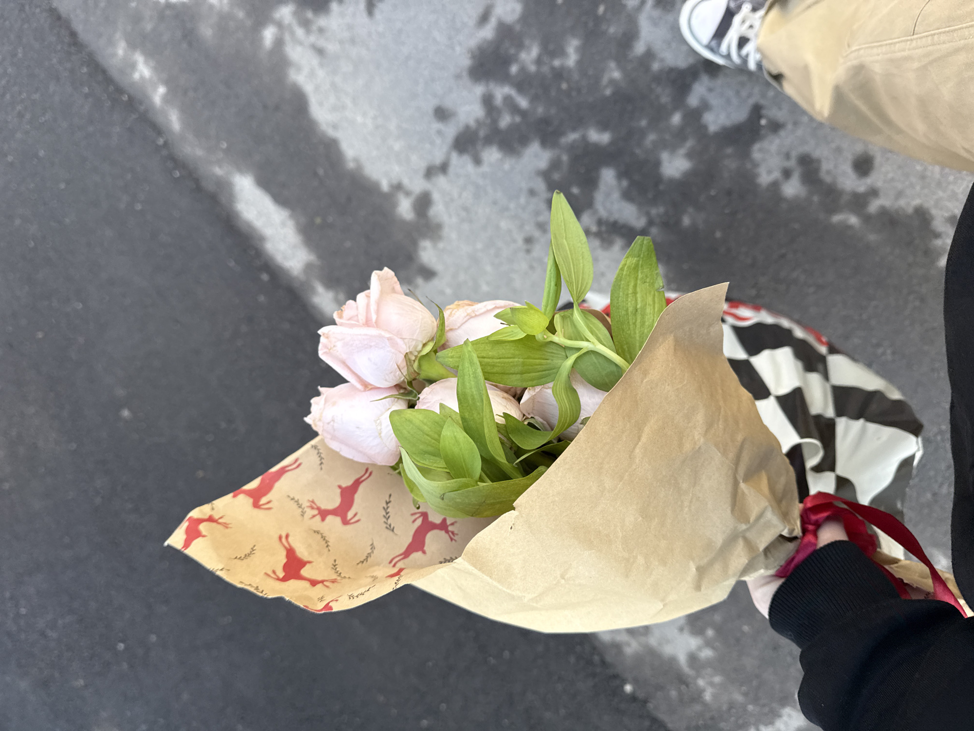 A person in tan pants and black and white sneakers stands on a city street in Yerevan, Armenia, holding a bouquet of light pink flowers wrapped in brown paper decorated with red reindeer. The bouquet is secured with a red ribbon. The person also carries a black and white checkered bag. The street appears wet. The person wears a black jacket.