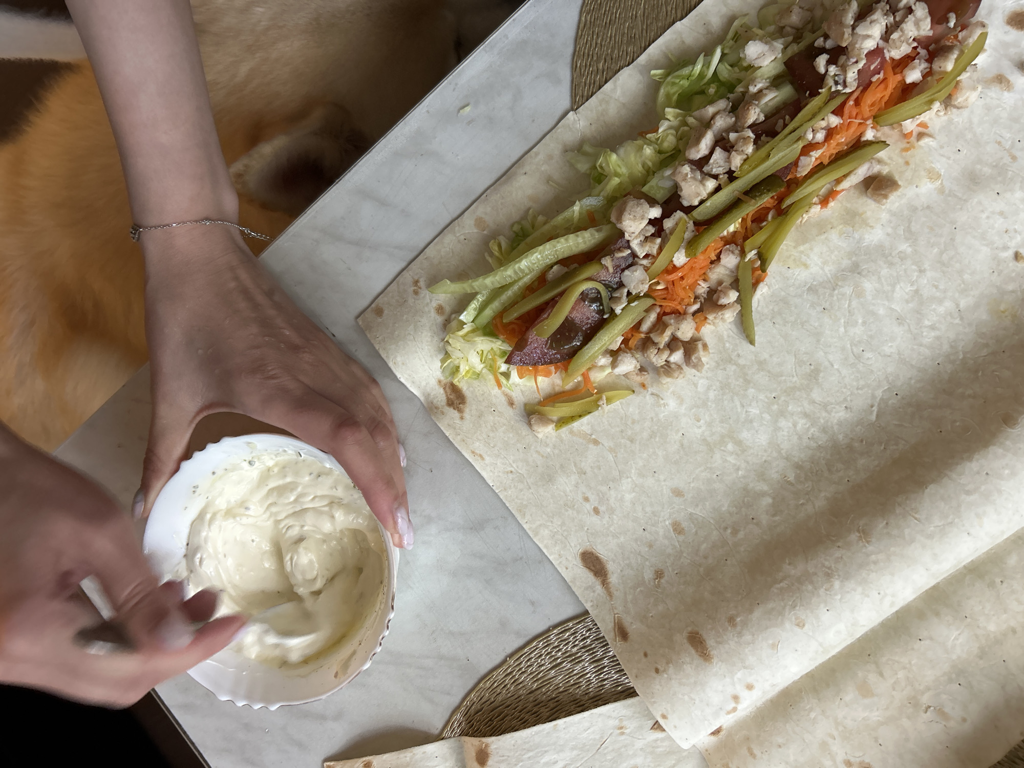 A person is preparing a shawarma. The scene is an indoor setting, likely a kitchen, with a marble countertop. The person's hands are visible, one holding a spoon and mixing a creamy white sauce in a small white bowl, while the other is near a rolled flatbread filled with ingredients. The flatbread contains shredded lettuce, shredded carrots, chopped chicken, and sliced pickles. A person's arm wearing a delicate silver bracelet is also in view. The lighting suggests daytime. There is no visible text in the image.