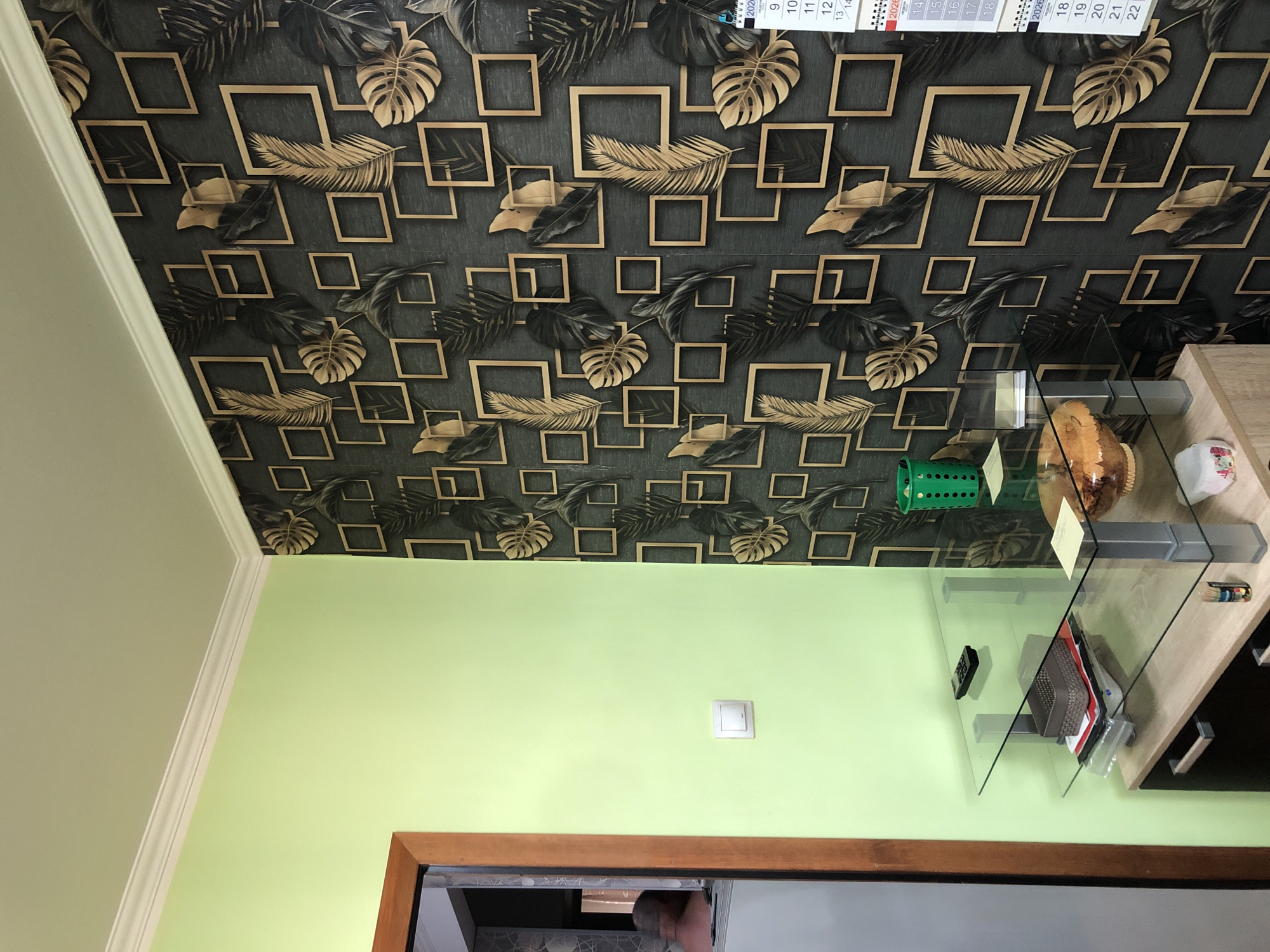 The image shows an interior scene, likely a room in a residential or commercial building. The walls are painted a light green on the left and covered with dark wallpaper featuring a geometric pattern of gold squares and tropical leaves on the right. A white electrical outlet is visible on the green wall. In the foreground, there is a modern glass-top shelf unit with a light wood and dark cabinet base. On the shelves, there are various items including a remote control, a woven basket, some papers, and a bowl with a pastry inside. A green trash can is also on the right shelf. On the right side of the image, a calendar from 2020 is attached to the wallpaper. A doorway with a wooden frame is visible on the far left, revealing a glimpse of another room or hallway with a person partially visible. The lighting appears to be artificial, and the overall impression is of a well-lit indoor space. The image does not provide any explicit visual cues that would definitively place it in Safonovo, Russia, or any specific city.