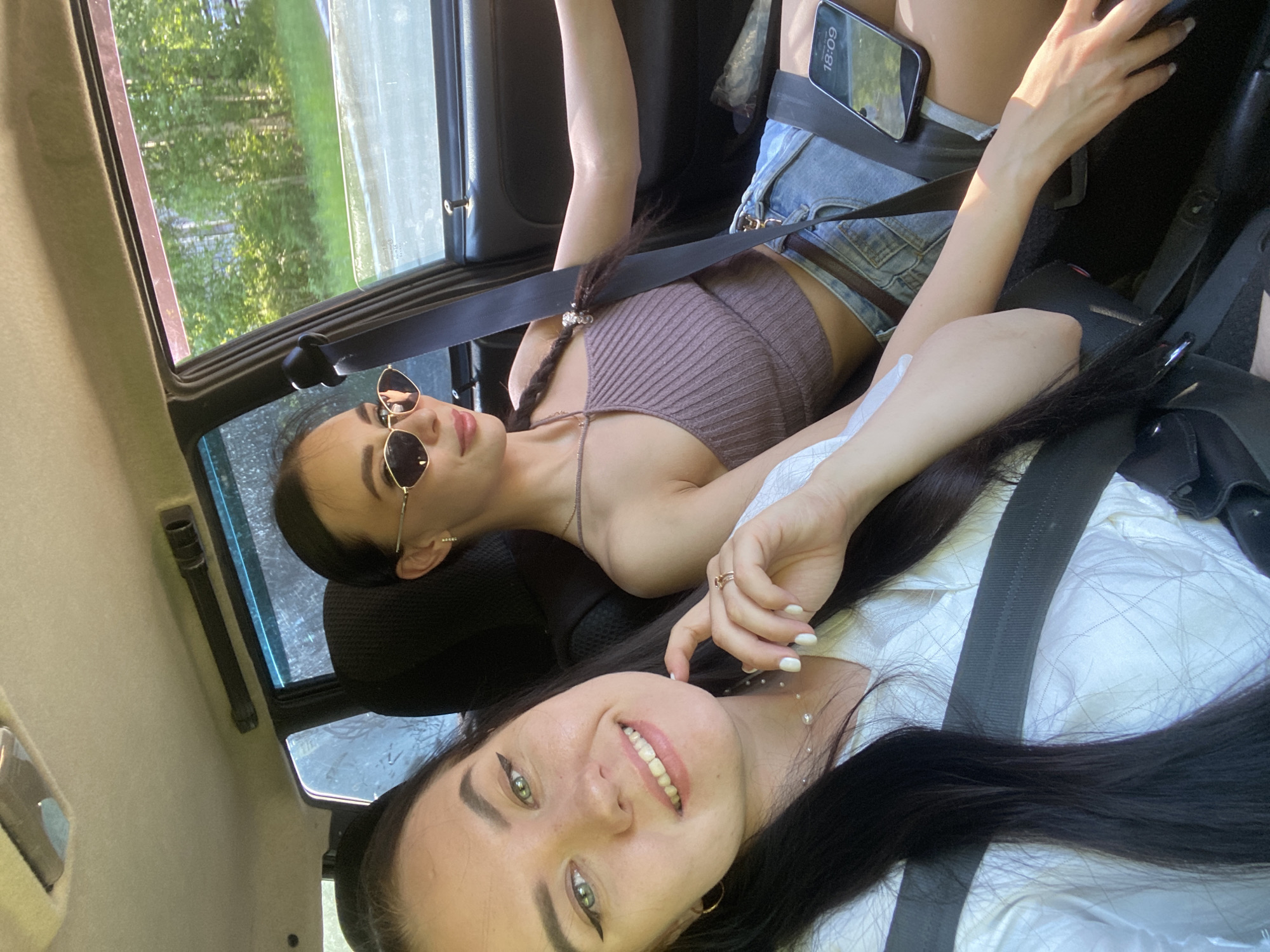 Two young women are in a car, both wearing seatbelts. The woman in the foreground is smiling, with long, dark hair, light eyes, and wearing a white patterned shirt. She has a ring on her finger and a necklace. The woman behind her is wearing sunglasses and a brown halter top with denim shorts, her hair is in a braid, and a phone is visible on her lap. They are presumably traveling, as a green, forested area is visible through the car window. The location is Chaikovsky, Russia.