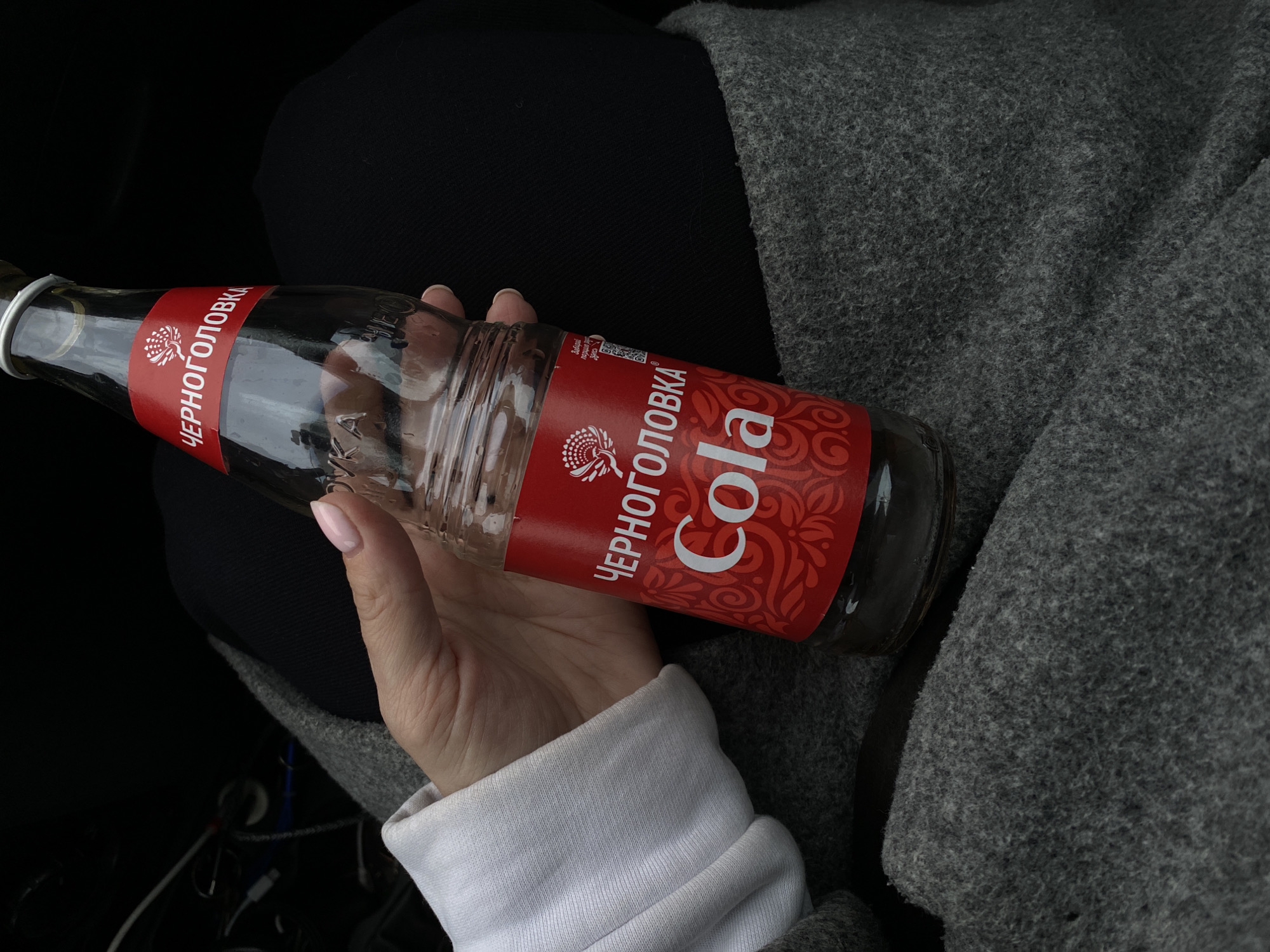 A glass bottle of "Chernogolovka Cola" is held in a woman's hand. The bottle is mostly empty, with traces of liquid at the bottom. The label is red with white Cyrillic text ("Черноголовка") and "Cola" written in English. The woman is wearing a white long-sleeved shirt, black pants, and a gray coat. The photograph was likely taken indoors, possibly in a car. There is no explicit evidence the location is Chaikovsky, Russia.