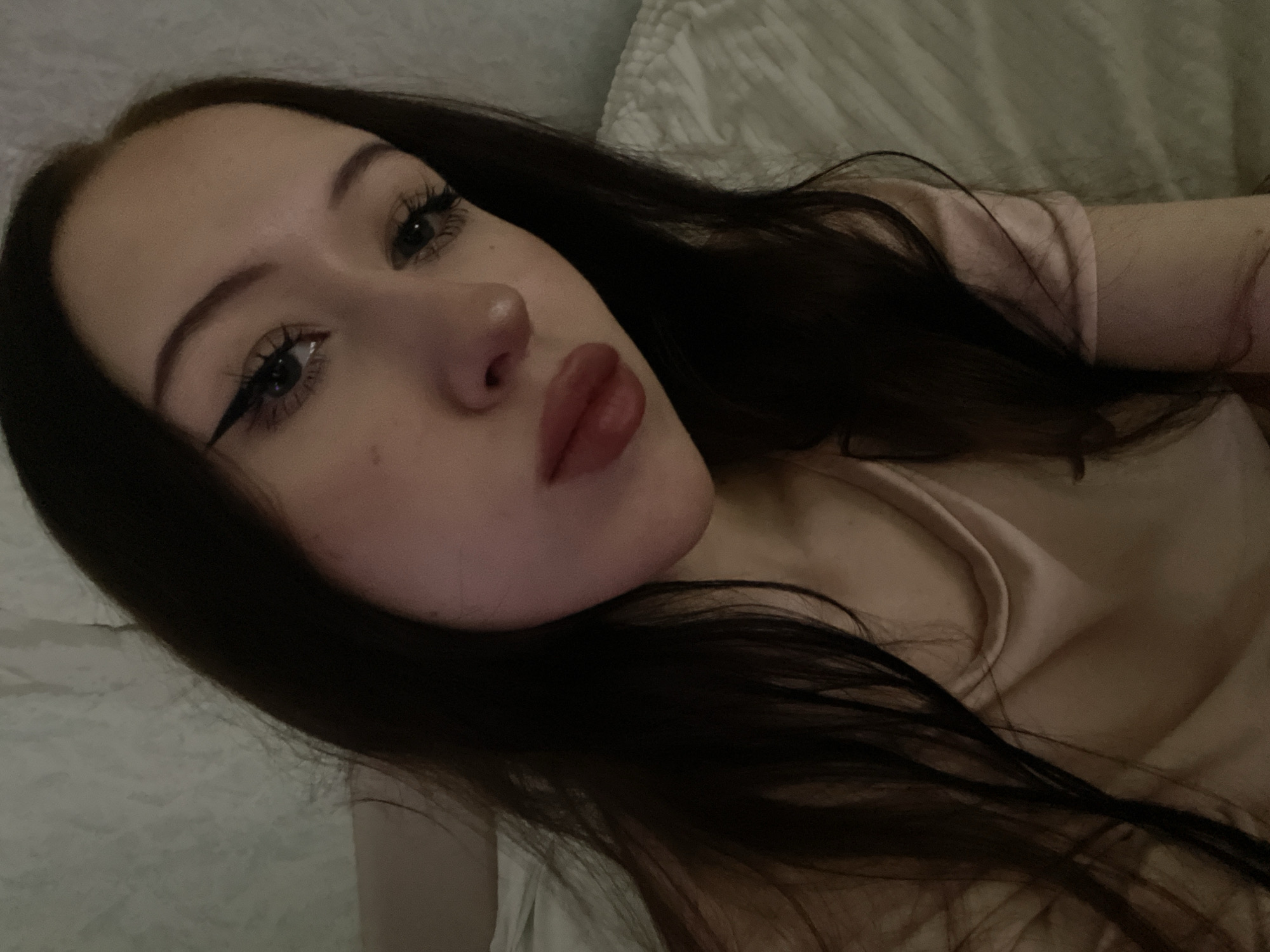 A medium shot photograph shows a young woman from the chest up, angled slightly to her right. She has long, dark brown hair that falls over her shoulders and down her chest. Her skin is fair, with visible makeup including winged eyeliner and mascara on her eyelashes, and lipstick on her lips. The background is out of focus, appearing to be a bed with a textured surface, possibly a headboard. The lighting is dim, casting a soft glow on her face and suggesting an indoor setting. The photograph was taken in Votkinsk, Russia.