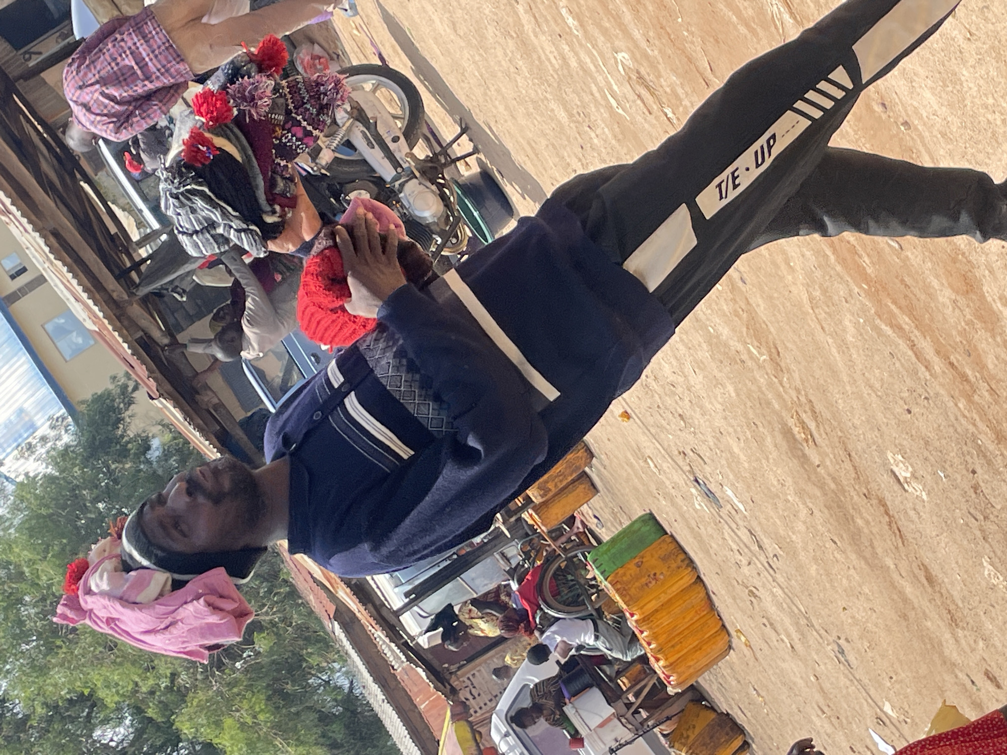 The image depicts a man in Wang, Nigeria, carrying a load of clothing on his head. He is wearing a navy blue long-sleeved shirt with white stripes and black track pants with white accents. He is holding a red knitted cap in his hands. In the background, there is a market scene with several stalls, a motorcycle loaded with clothing, and other people. The ground is dirt, and there are some yellow and green containers visible. The overall impression is of a busy marketplace, possibly in the late morning or early afternoon given the lighting.