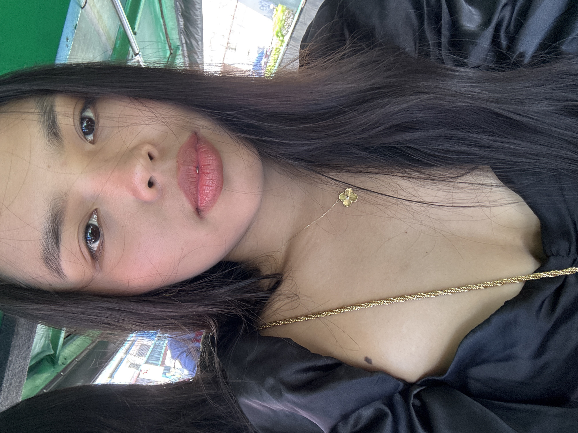 A close-up, tilted portrait captures a young woman, likely a selfie, with her gaze directed towards the camera. She has long, dark hair framing her face and shoulders. Her skin appears fair, with a slight flush on her cheeks, and her lips are painted a soft rosy pink. She wears a black, satin-like top or dress, and is adorned with two gold-colored necklaces: a delicate chain featuring a small, four-leaf clover pendant, and a thicker rope-style chain. A small mole is visible on her upper chest.
The background is blurred, showing an out-of-focus outdoor urban environment, possibly viewed from inside a vehicle or building in Davao City, Philippines. Glimpses of bright sky, multi-story buildings, and what appears to be part of a street scene are reflected in her eye and on metallic elements in the background. Green and silver metallic structures are also discernible in the soft-focus background, suggesting a vehicle interior or a specific architectural setting. The bright reflections indicate it is daytime. The woman's expression is neutral and calm. There is no visible text.