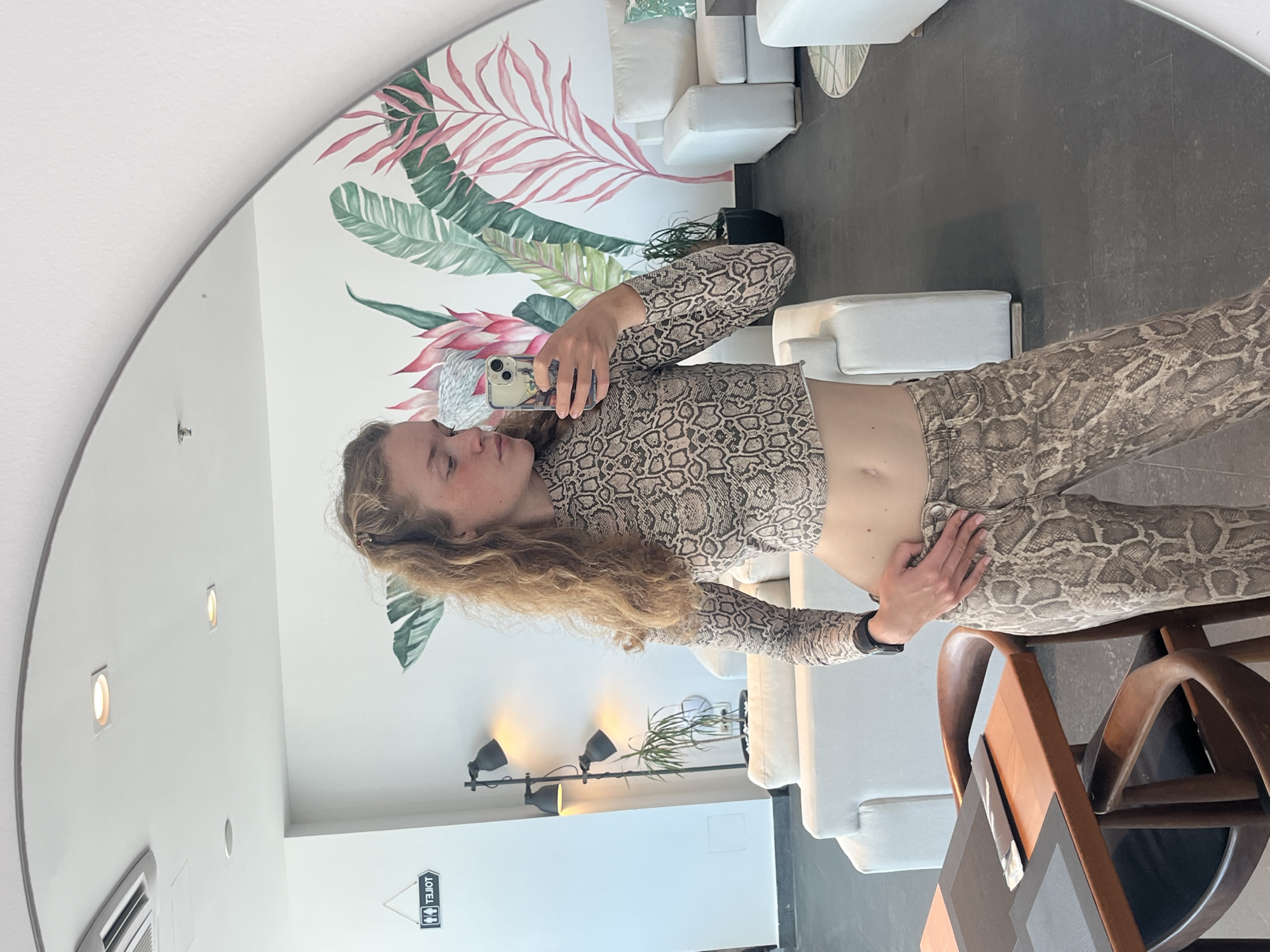 A woman is taking a mirror selfie indoors. She is wearing a matching snake print crop top and pants. The mirror is round and shows the reflection of a room with white walls and seating, a decorative wall mural with botanical illustrations, and various light fixtures. A toilet sign is visible on the wall. The location is likely within a business in Budva, Montenegro, based on the style and context.