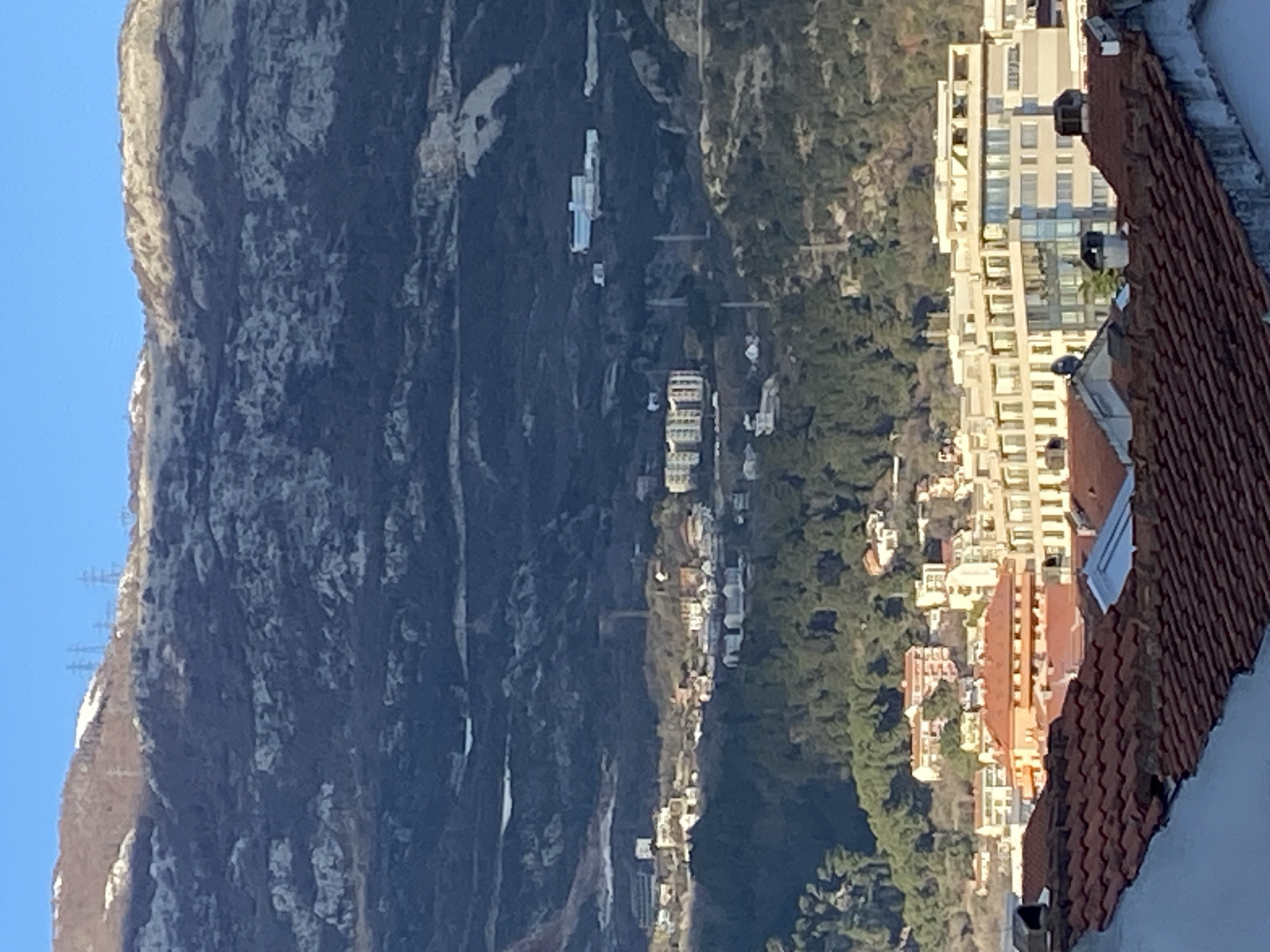 The image shows a view from a building in Budva, Montenegro, overlooking a landscape including a rocky mountain, dense vegetation, and buildings. The mountain occupies the left side of the frame, with visible variations in texture and color on its surface, and power lines are present near its peak. A line of buildings follows a path adjacent to the mountainside. A mix of residential and commercial buildings is visible, some with red tile roofs. On the right side of the frame, a red-tiled roof occupies a significant portion of the foreground. The sky is a clear blue in the upper left corner. The city of Budva is evident through the density and arrangement of the buildings and the overall urban layout.