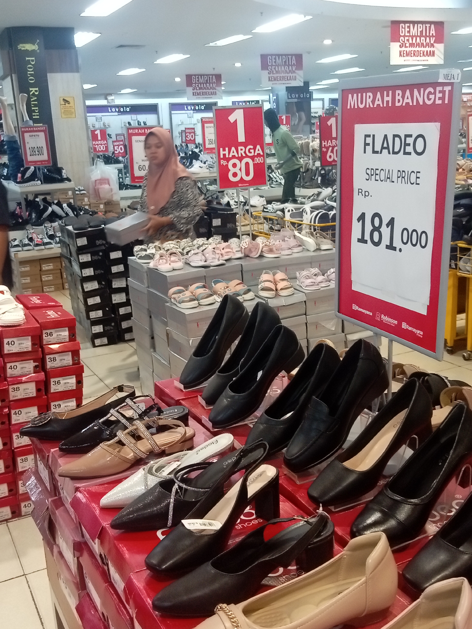 This is a medium shot, eye-level interior photograph of a shoe store in Batam, Indonesia, likely within a shopping mall or department store. The scene is brightly lit with fluorescent lighting, typical of retail environments. In the foreground, a display of women's shoes is arranged on top of stacked shoe boxes, predominantly black and neutral tones, with some embellished with rhinestones. The shoe boxes are stacked in a retail display format, some showing size labels like "36", "38", and "40". To the left, more shoe boxes are stacked, with a few pairs of sneakers visible on shelves above. A prominent red and white sign in the center right announces a sale for "FLADEO SPECIAL PRICE Rp. 181.000". Another sign in the background, also red and white, displays "MURAH BANGET" (very cheap). In the middle ground, a woman wearing a hijab and a patterned dress is holding a shoebox and appears to be interacting with the merchandise. Behind her, a man in a green shirt is also visible, seemingly working in the store. Several displays of shoes, including children's sandals, are visible further back, arranged on shelves and in boxes. In the far background, store signage includes "Lavola" and banners reading "GEMPITA SEMARAK KEMERDEKAAN" (Festive Independence Day). The overall impression is a busy retail environment during a promotional event.