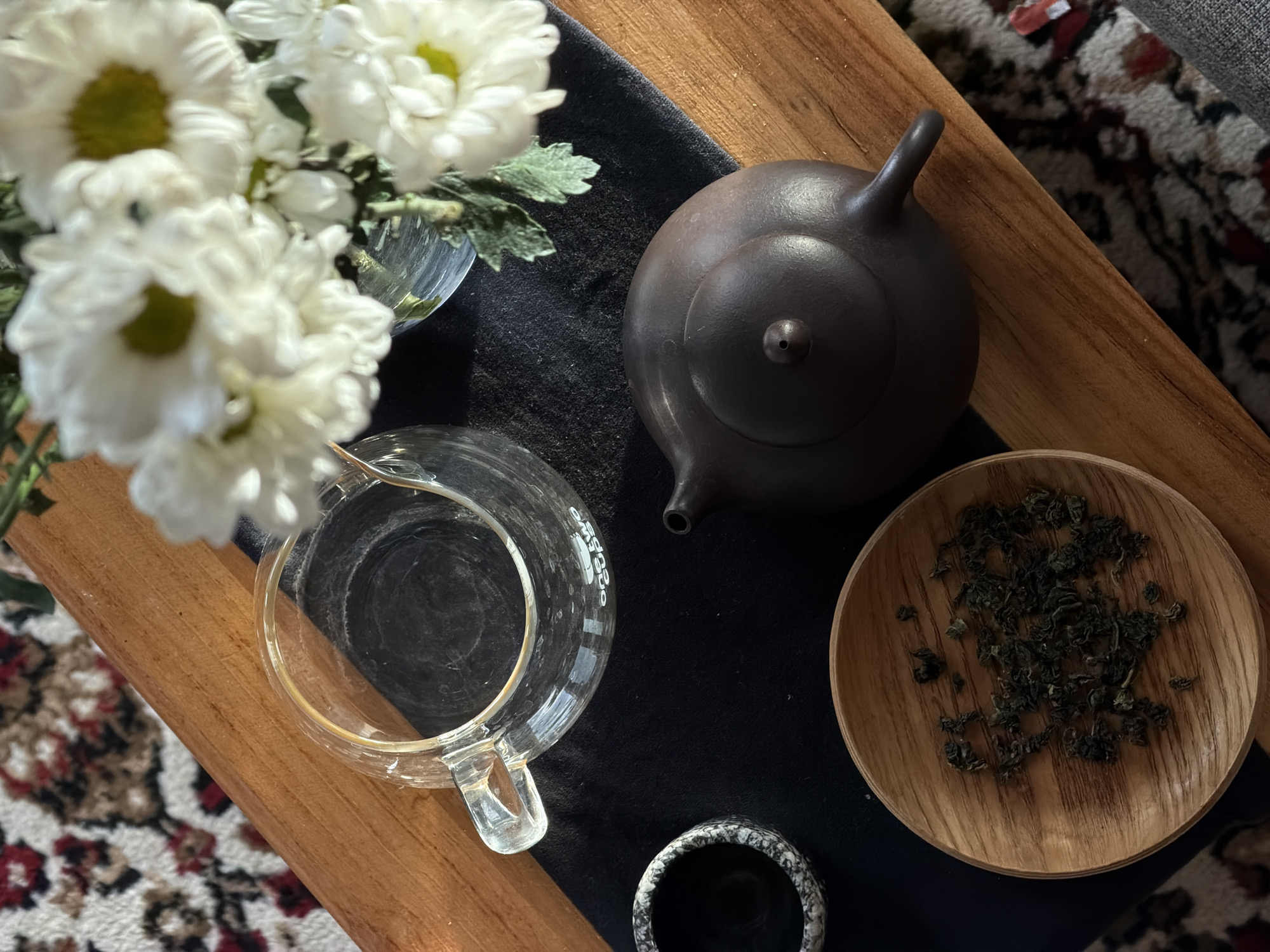 The image depicts a tea setting placed on a wooden surface, likely a small table or tray. A dark-colored teapot sits centered on a dark rectangular cloth, next to a small wooden plate filled with loose tea leaves. To the left is a clear glass pitcher with a handle. A small vase containing white flowers with green centers is placed to the upper left. A small, mottled cup sits at the lower center. The background includes a patterned rug. No people are visible. There is no information within the image to confirm this scene is located in North Kuta, Indonesia.