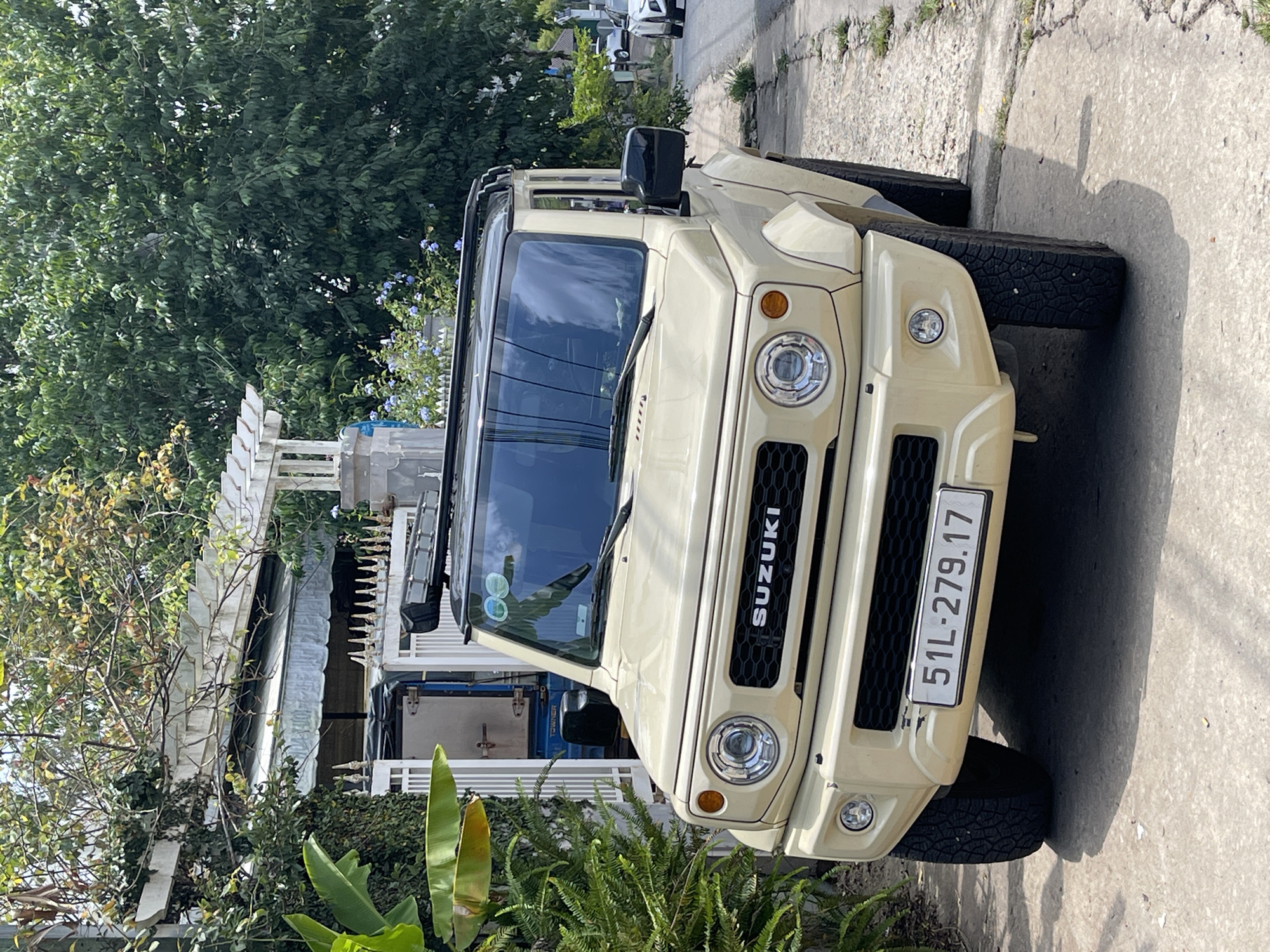 A front-view shot of a tan Suzuki Jimny is on a concrete road in a residential area in Cam Ly - Đà Lạt, Vietnam. The license plate is visible, reading "51L-279.17." The Jimny sits in front of a white building partially obscured by green foliage. More greenery is visible in the foreground. Other cars are parked further up the road in the distance. The time of day is daytime, with sunlight casting shadows on the road.