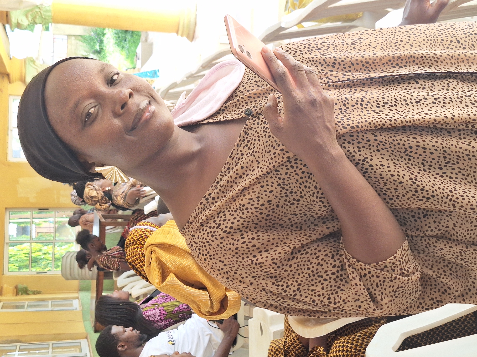 The image shows a Black woman in Rido, Nigeria, wearing a black head covering and a beige and brown patterned dress. She is holding a peach-colored smartphone in her right hand. Behind her, a group of people are seated, some wearing patterned clothing. A yellow fabric is draped near the woman. Windows are visible in the background, with trees and greenery outside. The setting appears to be indoors, with white chairs and yellow walls.