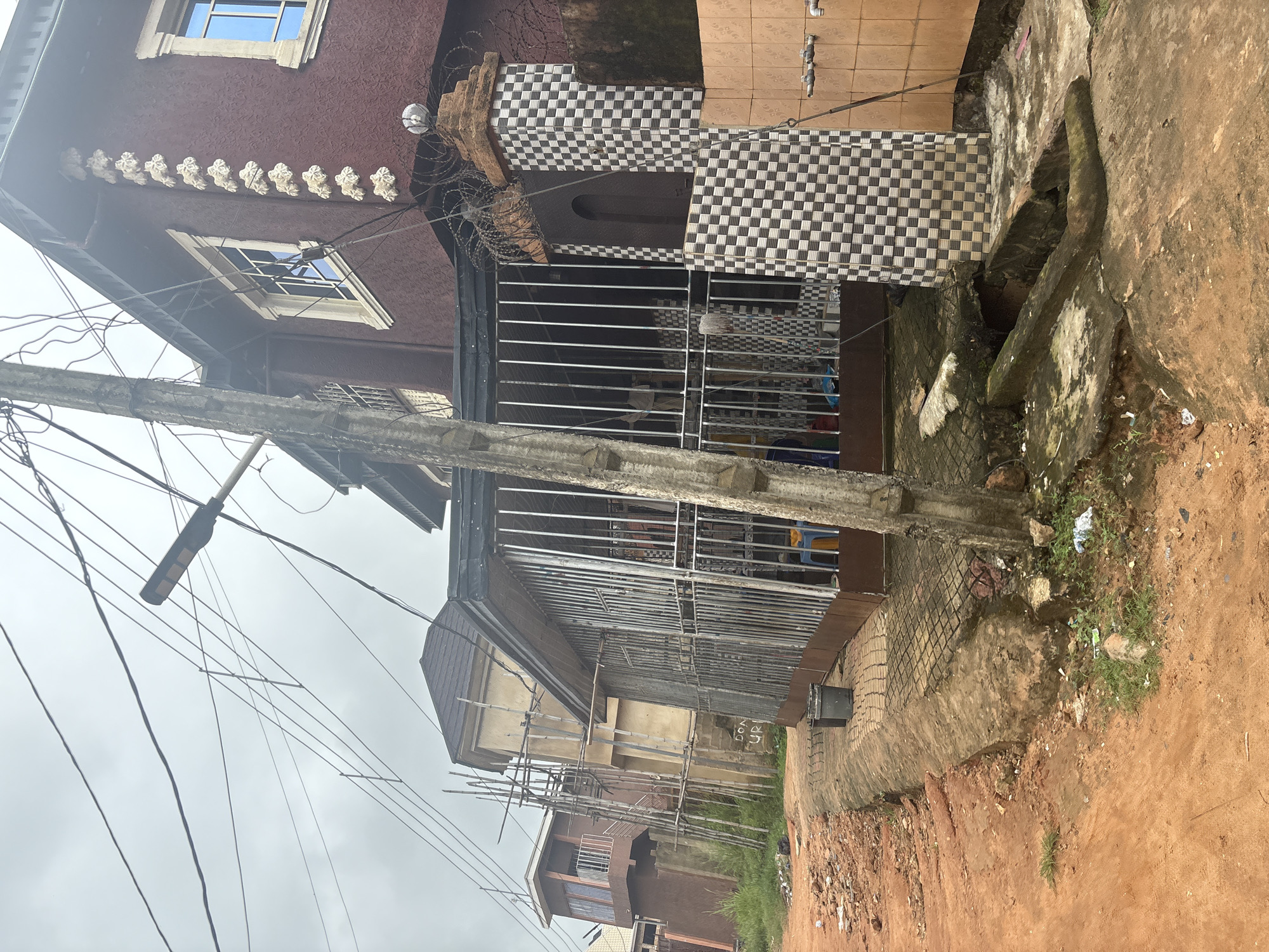 The image depicts a street scene in Obosi, Nigeria, with a focus on residential buildings and infrastructure. The foreground shows a reddish-brown dirt road with debris and patchy grass. To the right, a concrete walkway leads to a house with a gated veranda. The house is painted maroon and features decorative textured walls and white-trimmed windows. A checkered tiled pillar stands beside the entrance, topped with barbed wire and a decorative light fixture. Adjacent to this pillar is a section of tiled wall with two faucets.
Behind the foreground building, other structures are visible. One building appears to be under construction, with bamboo scaffolding. Another building, further back and to the left, has a balcony with railings. A prominent concrete pole with numerous electrical wires stretches across the scene, indicating the presence of overhead power lines. The sky is overcast and gray, suggesting a cloudy day. The overall impression is of a developing urban or semi-urban area with a mix of completed and unfinished constructions, common utilities, and unpaved roadways.